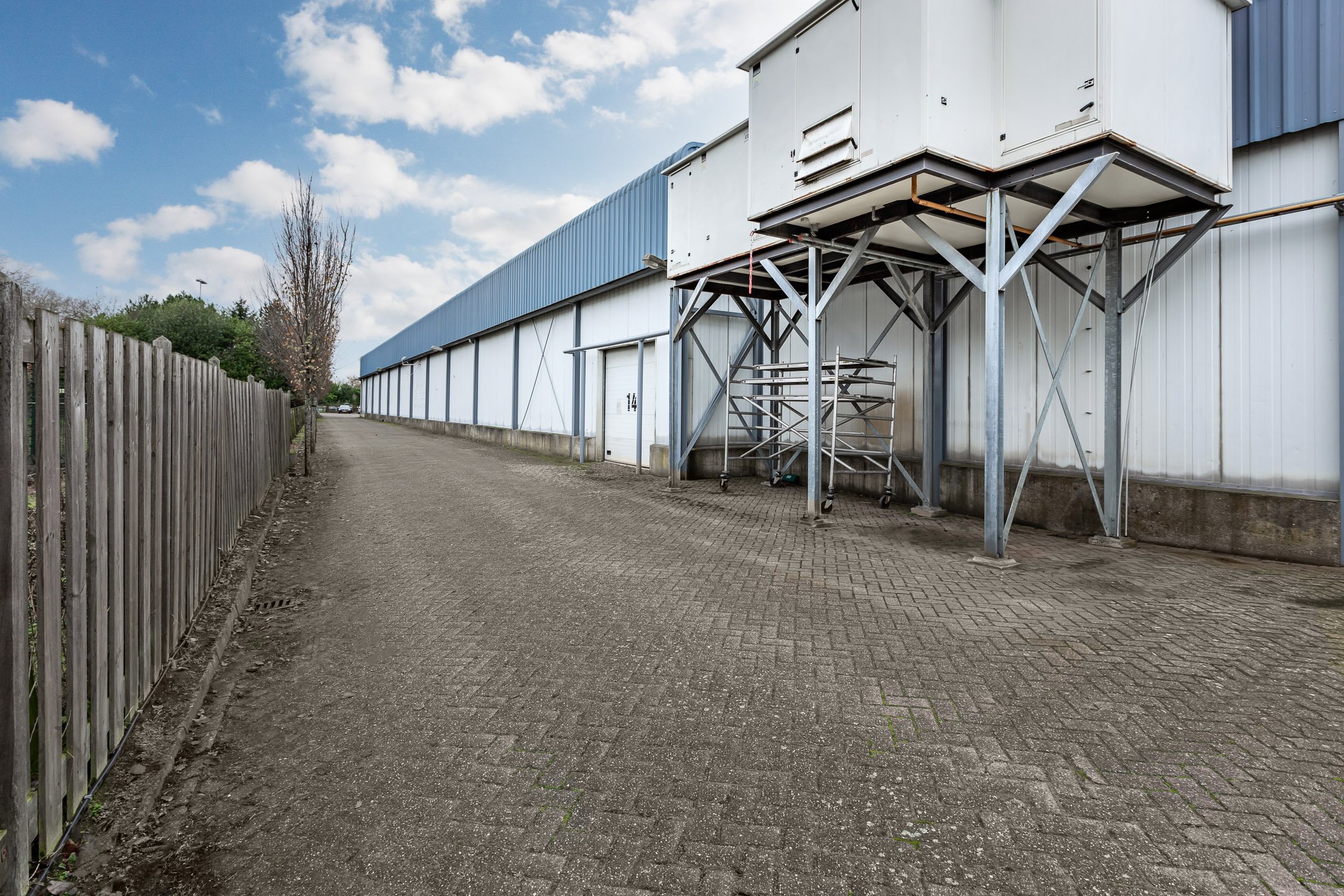 Empty alleyway beside a large industrial building with metal exterior walls and a blue roof, featuring a fenced side and some industrial equipment near the wall.