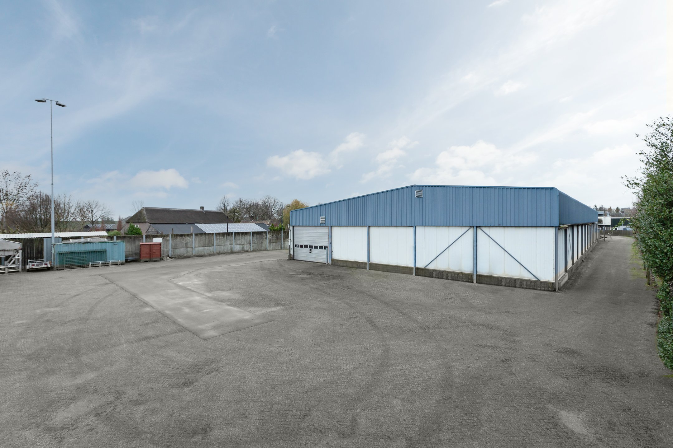 Empty parking lot with a large industrial blue and white building, fence, and a few small trees, under a partly cloudy sky.