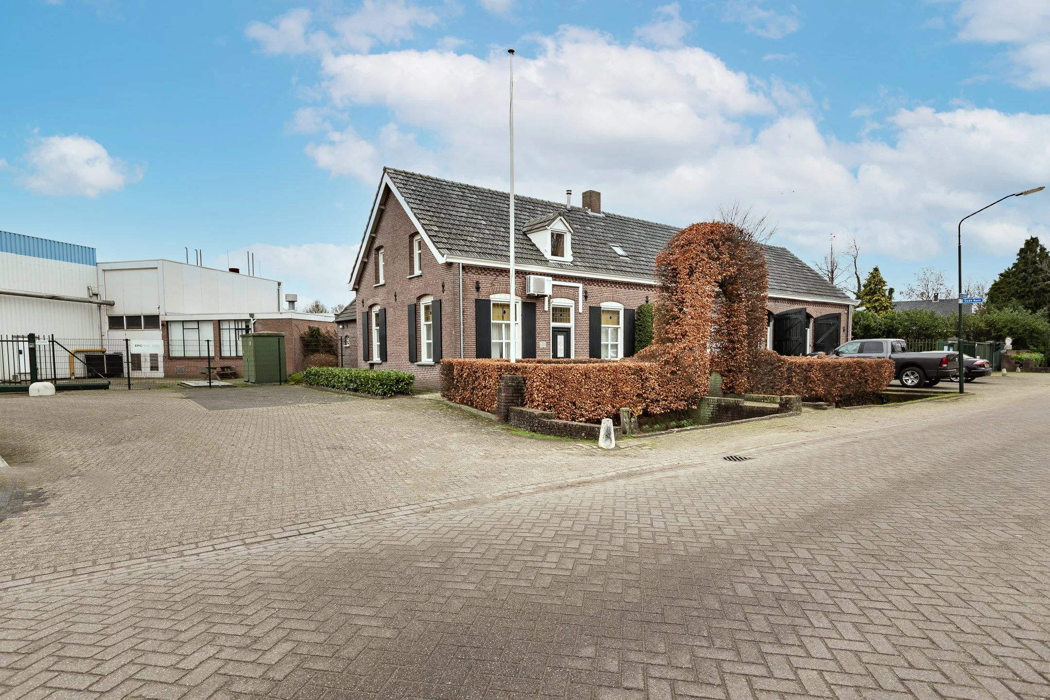 A brick house with a sloped roof, several windows, a chimney, and a hedge in front. The house has an air conditioning unit and a black garage door. There are cars parked, a street sign, streetlight, and industrial buildings nearby, under a partly cloudy sky.