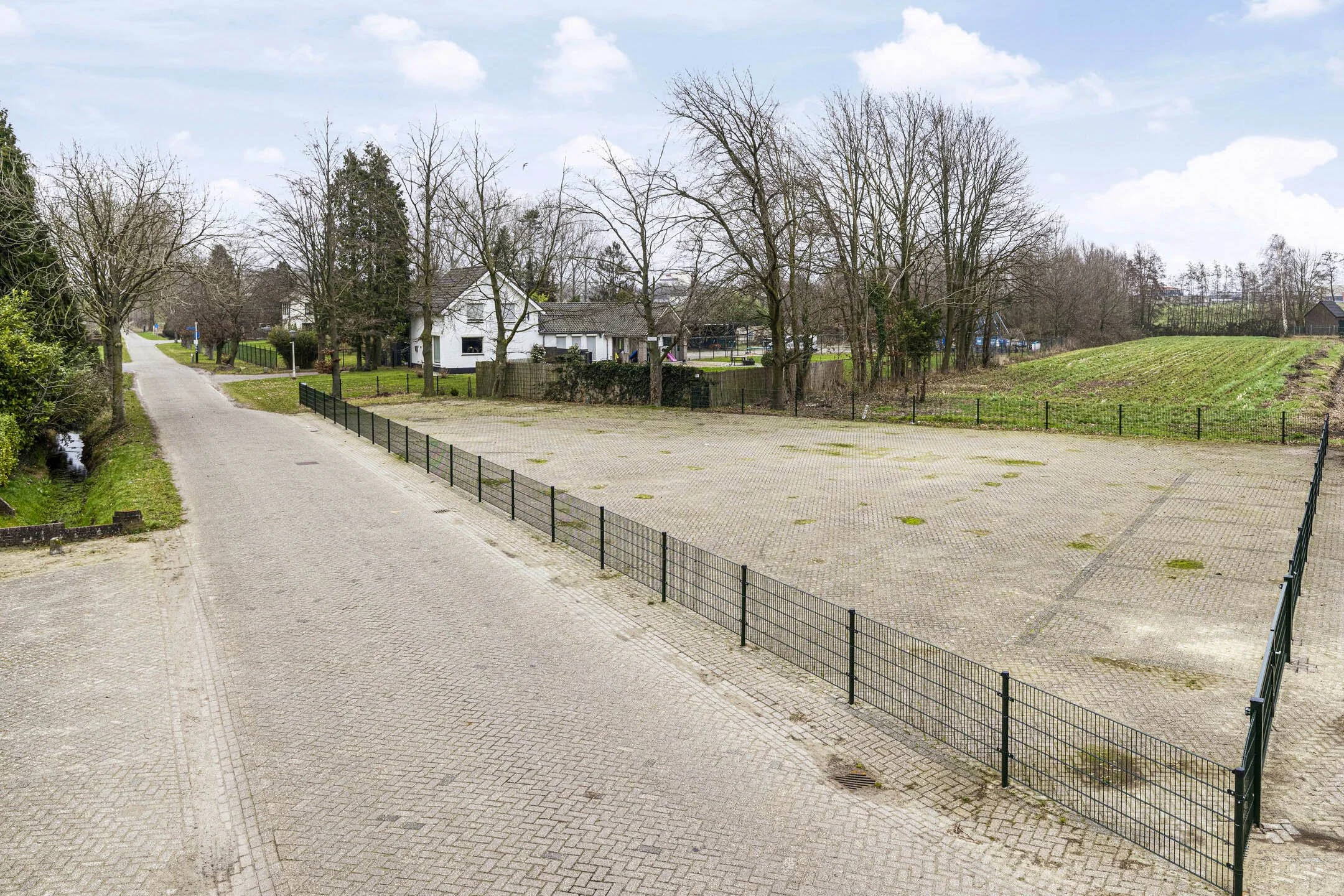 Empty paved outdoor area with a small fenced section on the right, a house with trees in the background, and a pathway leading into a green field.