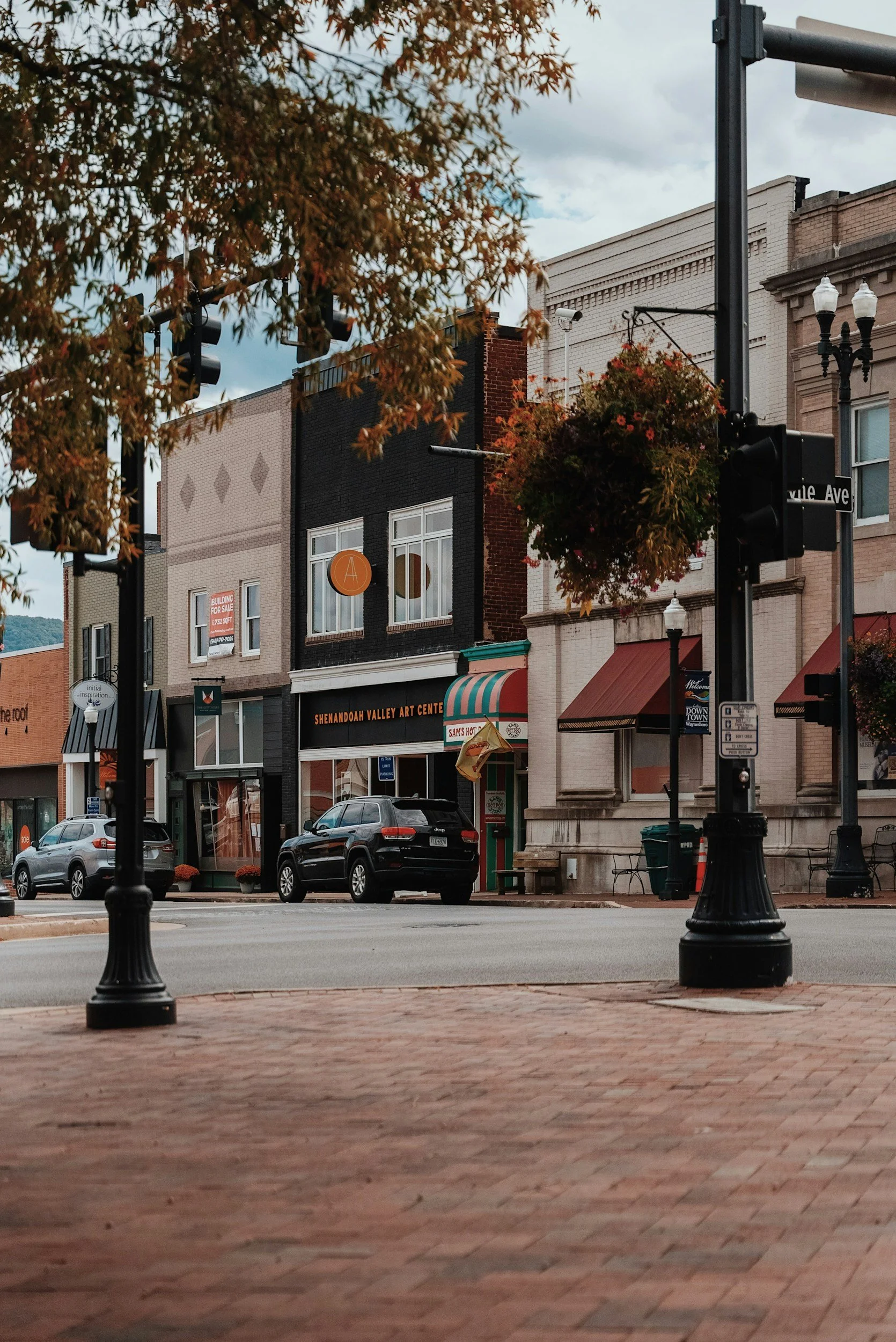 View of a downtown street with brick sidewalks, black street lamps, a few parked cars, and a row of buildings including an art center and storefronts, with a tree with reddish leaves in the foreground.