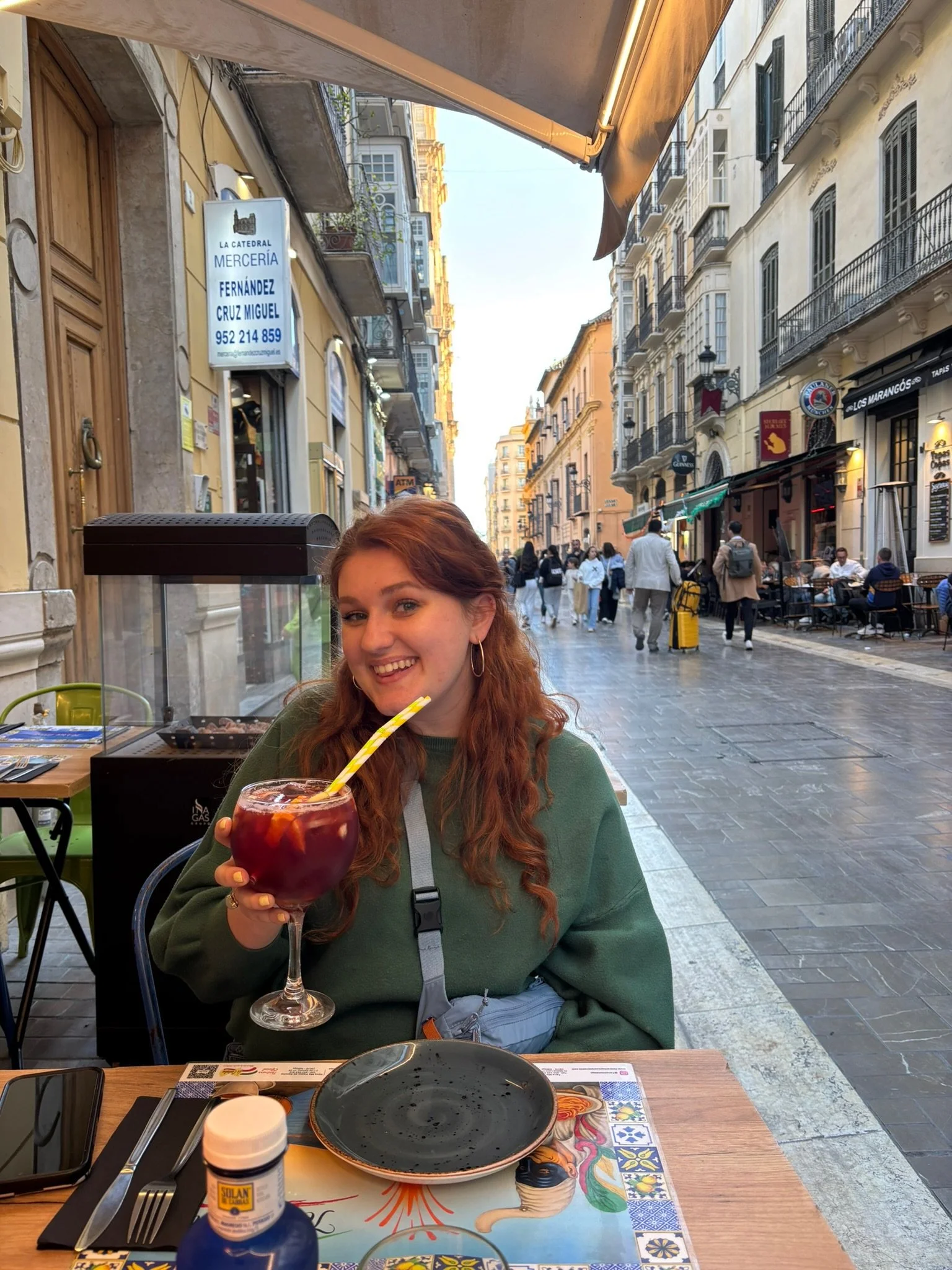 A smiling woman with long red hair sitting at an outdoor restaurant table, holding a glass of red sangria with fruit and a yellow striped straw. The table has a colorful placemat with a depiction of a mermaid, utensils, and an empty black plate. The street behind her is lively with people walking, shops, and cafes, with buildings featuring balconies and ornate architecture.