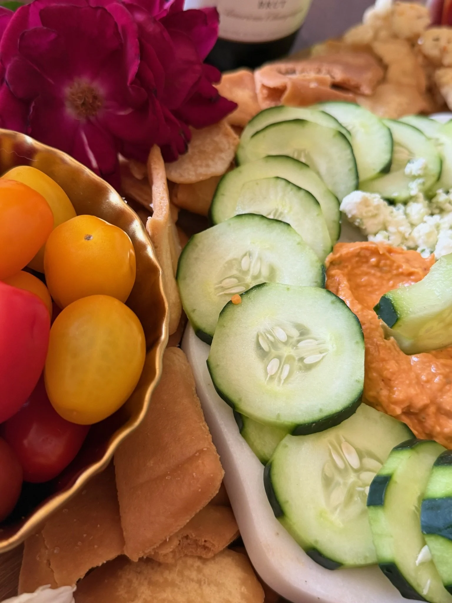 Close-up of a platter with cucumber slices, cherry tomatoes, and a scoop of guacamole, with a pink flower and other snacks in the background.