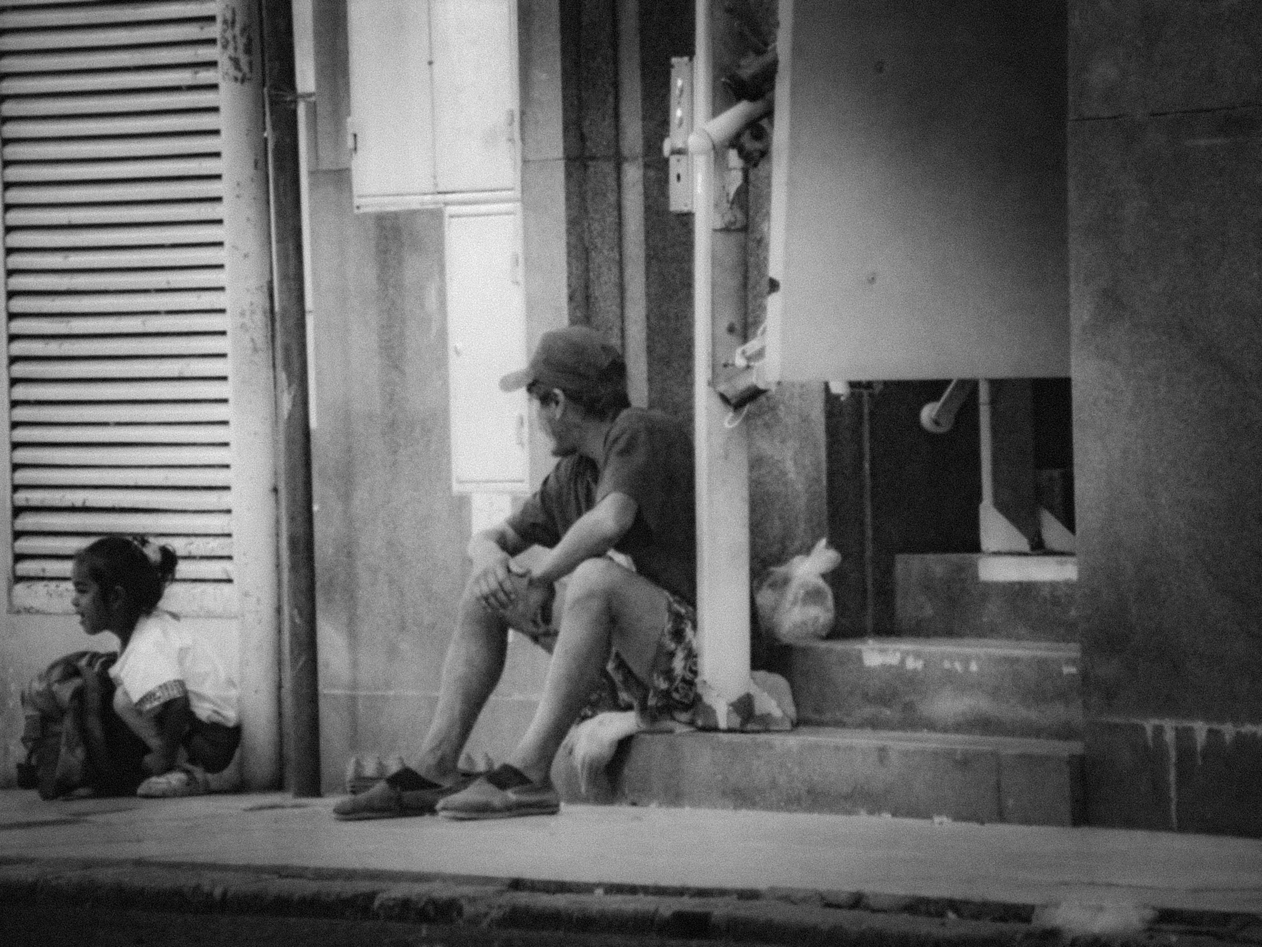 A person wearing a cap and shorts is lying on the floor with their eyes closed, appearing to rest or sleep. There are other children sitting against the wall in a classroom or waiting area.