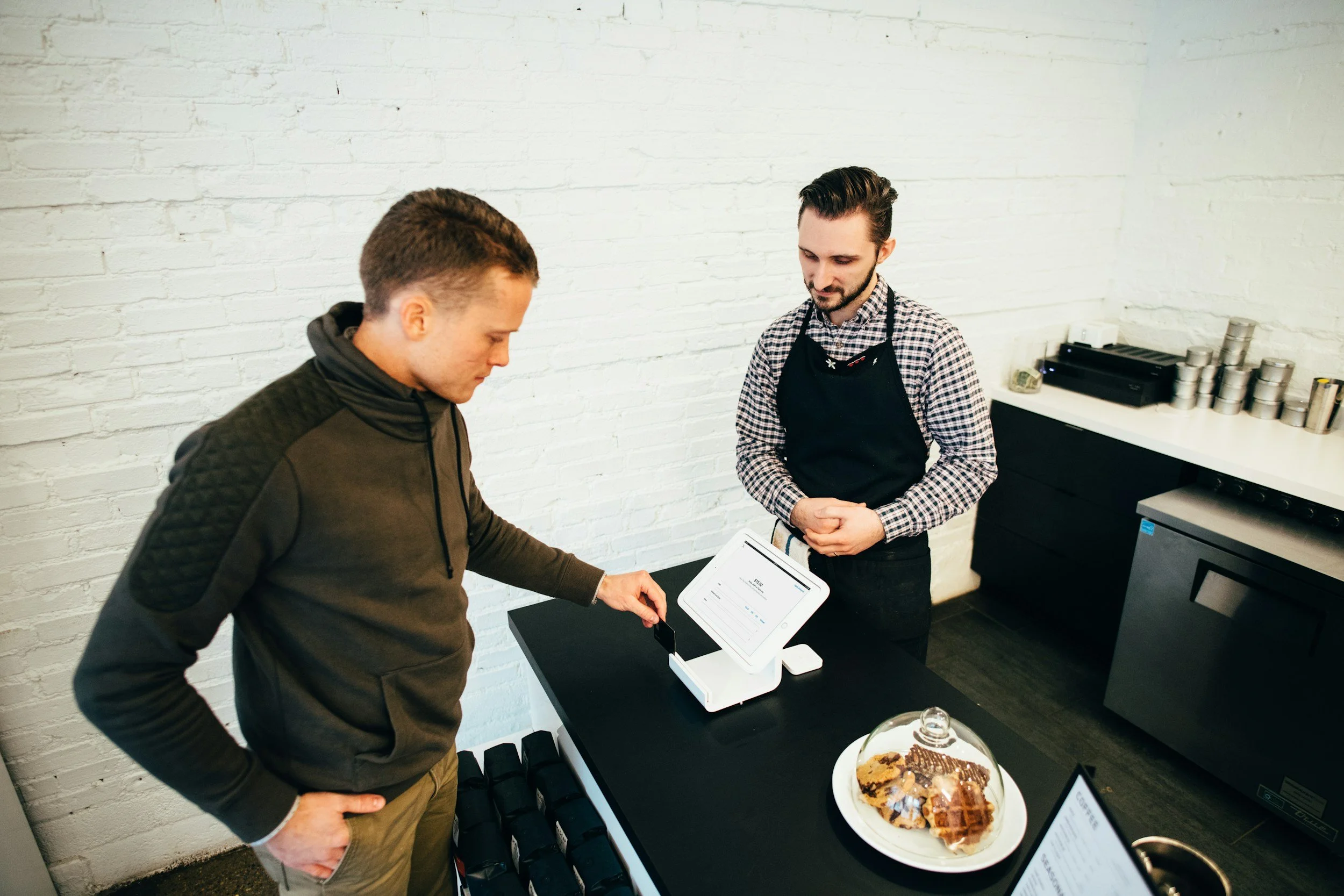 Two men are at a counter, one is using a touchscreen point of sale system while the other observes. A large plate with a dessert topped with chocolate sauce is on the table in front of them. The background features a white brick wall and some stacked cans and boxes.