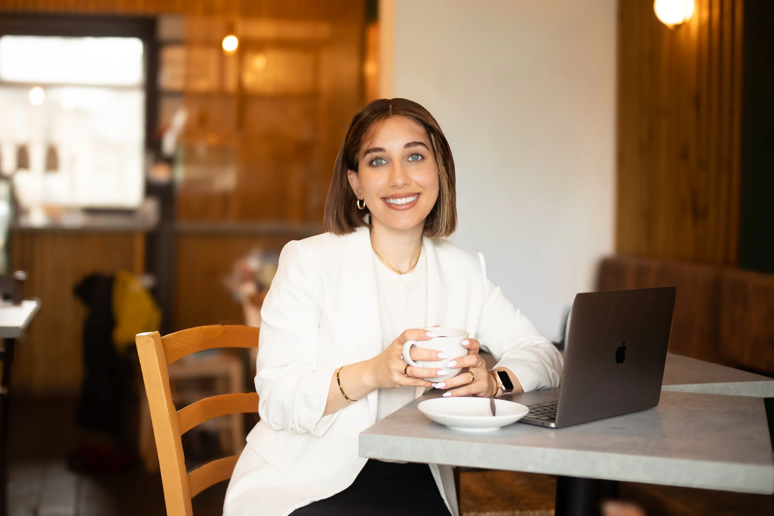 A woman with short brown hair and blue eyes sitting at a table in a coffee shop, smiling and holding a white coffee cup. She has a white blazer, gold jewelry, and is seated in front of a laptop. The background features warm wooden decor and soft lighting.