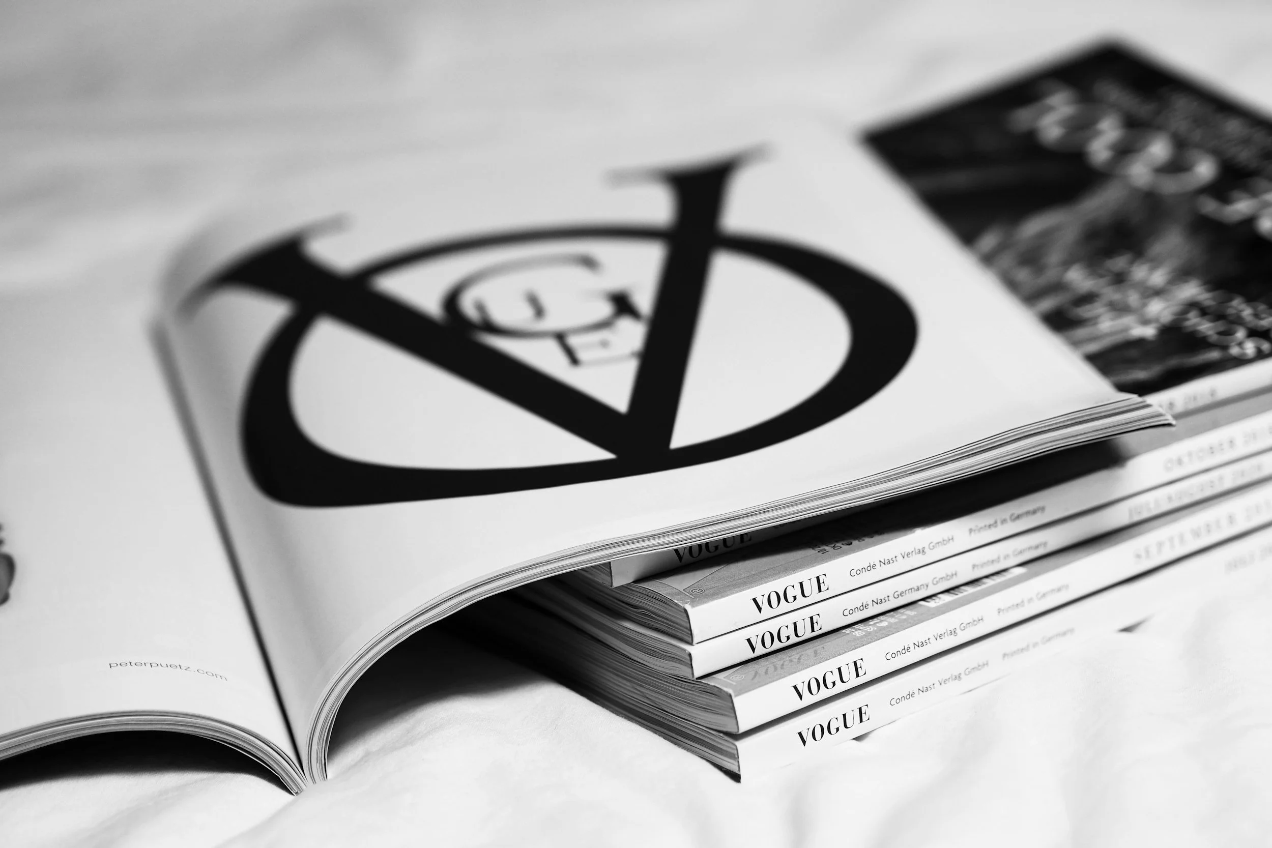 Black and white photo of open magazines stacked on a table, with the word 'VOGUE' visible on the spines.