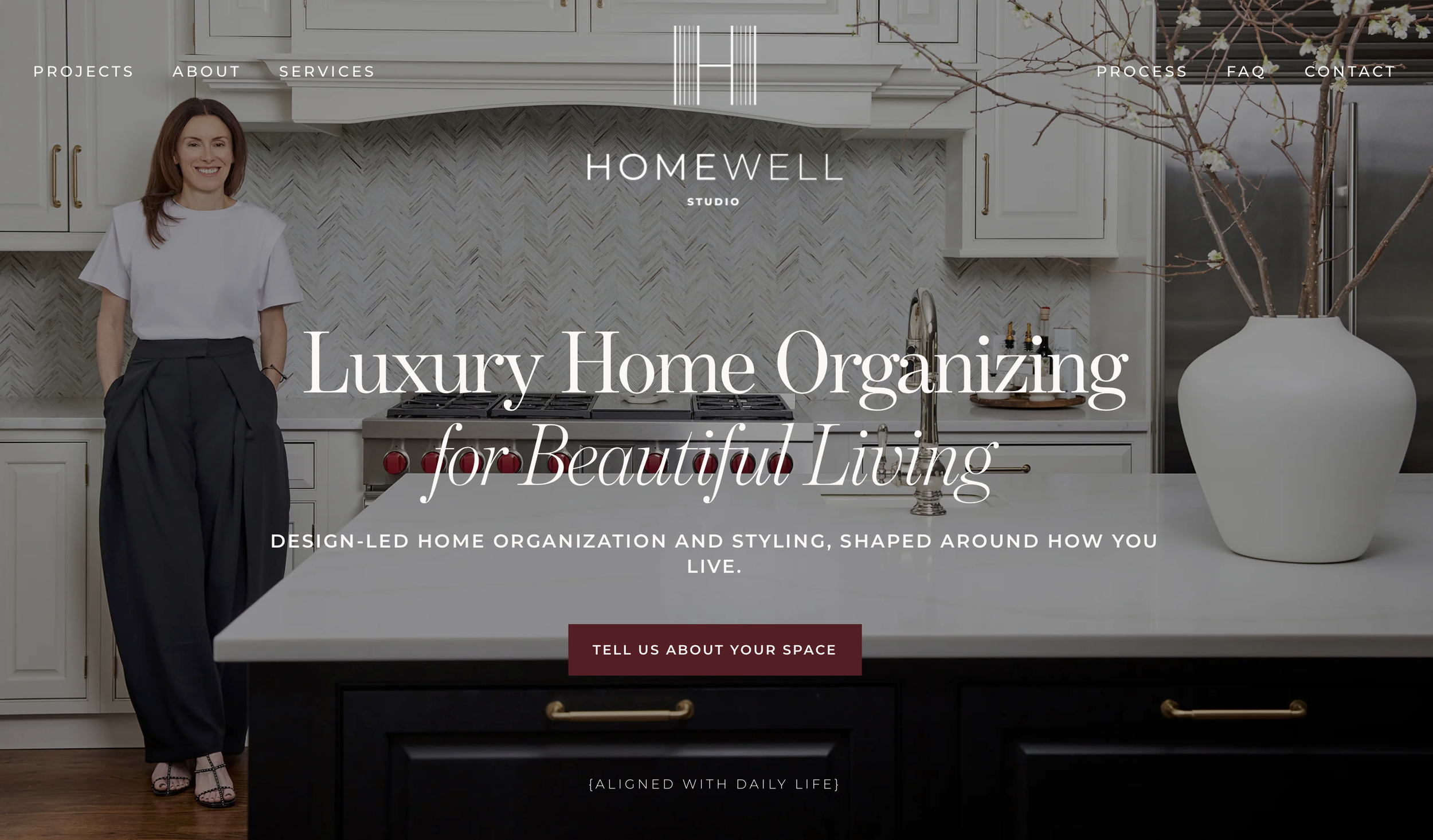The image shows a woman smiling and standing in a modern white kitchen with a large white vase on the countertop. The kitchen features white cabinets with gold handles, a gray patterned backsplash, and a stainless steel refrigerator. The website overlay includes navigation links and text about luxury home organizing and styling.