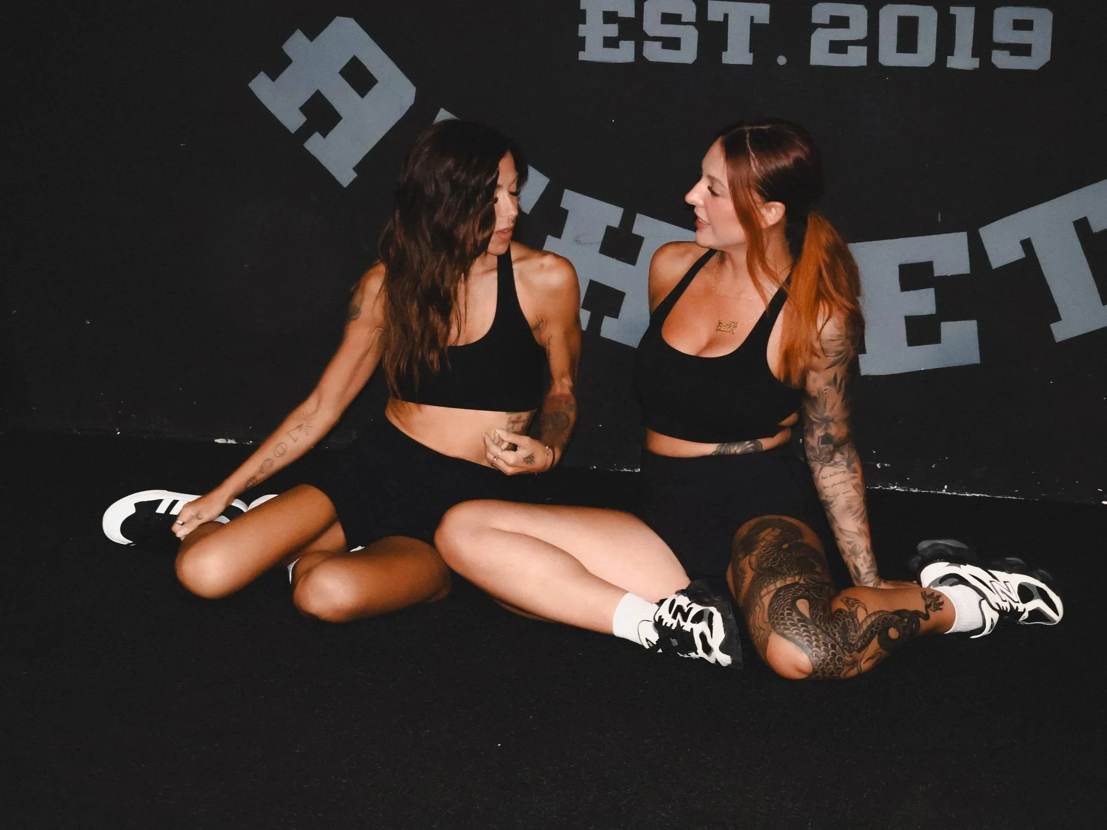 Two women sitting closely on the floor, wearing black sports bras and shorts, with tattoos on their arms and legs, and wearing sneakers, against a black wall background with white text.