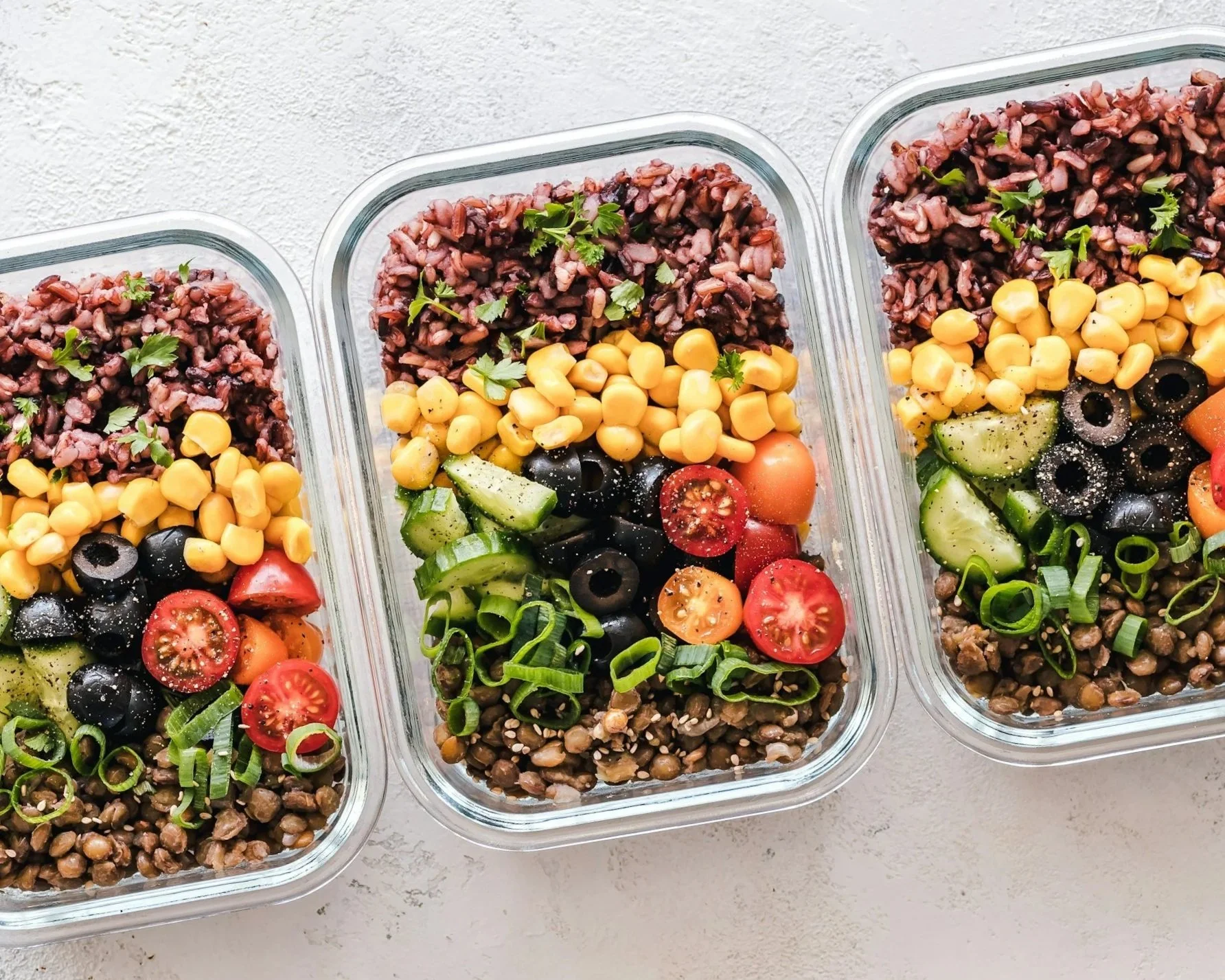 Three glass meal prep containers filled with a colorful salad including black beans, cherry tomatoes, sliced cucumbers, chopped green onions, corn, and cooked lentils. The salads are garnished with fresh parsley. The containers are placed on a light-colored surface.