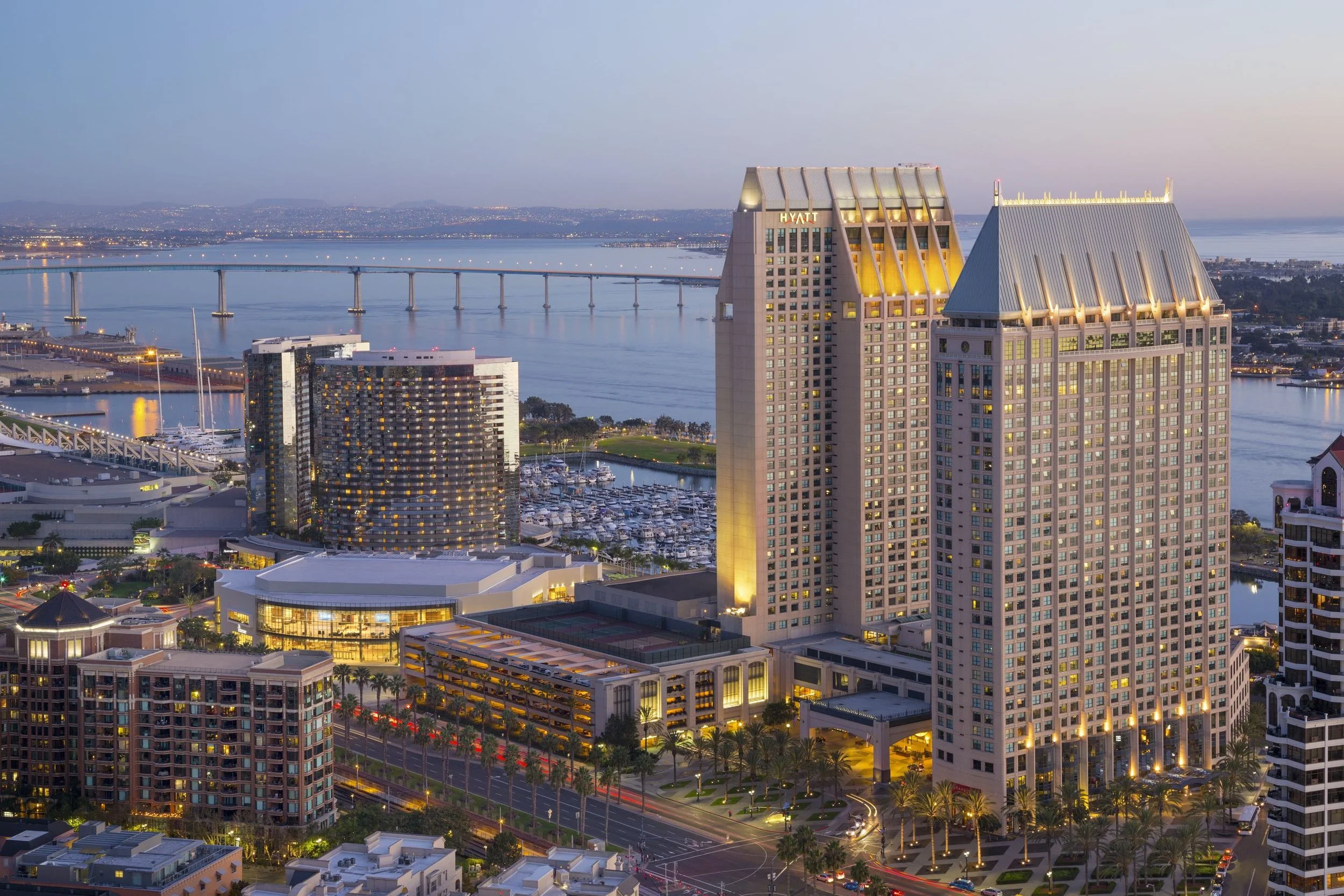 A cityscape at dusk featuring tall buildings, a bridge over a river, and a marina with boats. The buildings are illuminated, with one displaying the Hyatt logo.