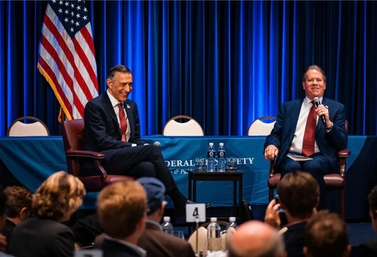 Two men in suits sitting on stage at a panel discussion, with an American flag and blue curtains behind them, audience members in front, and water bottles on a table between them.