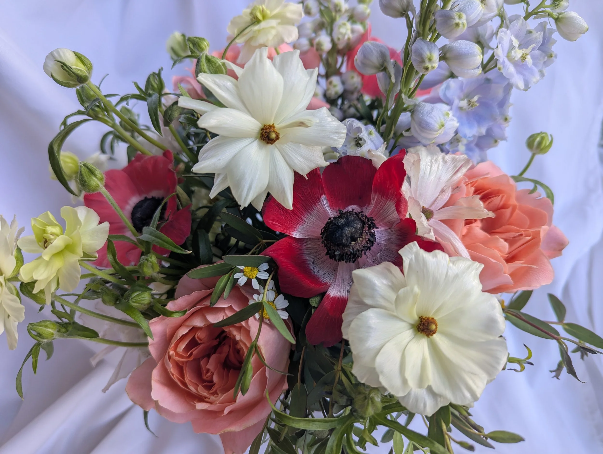 Close-up of a bouquet of mixed flowers, including white, pink, red, yellow, and blue flowers, with some buds and green leaves visible.