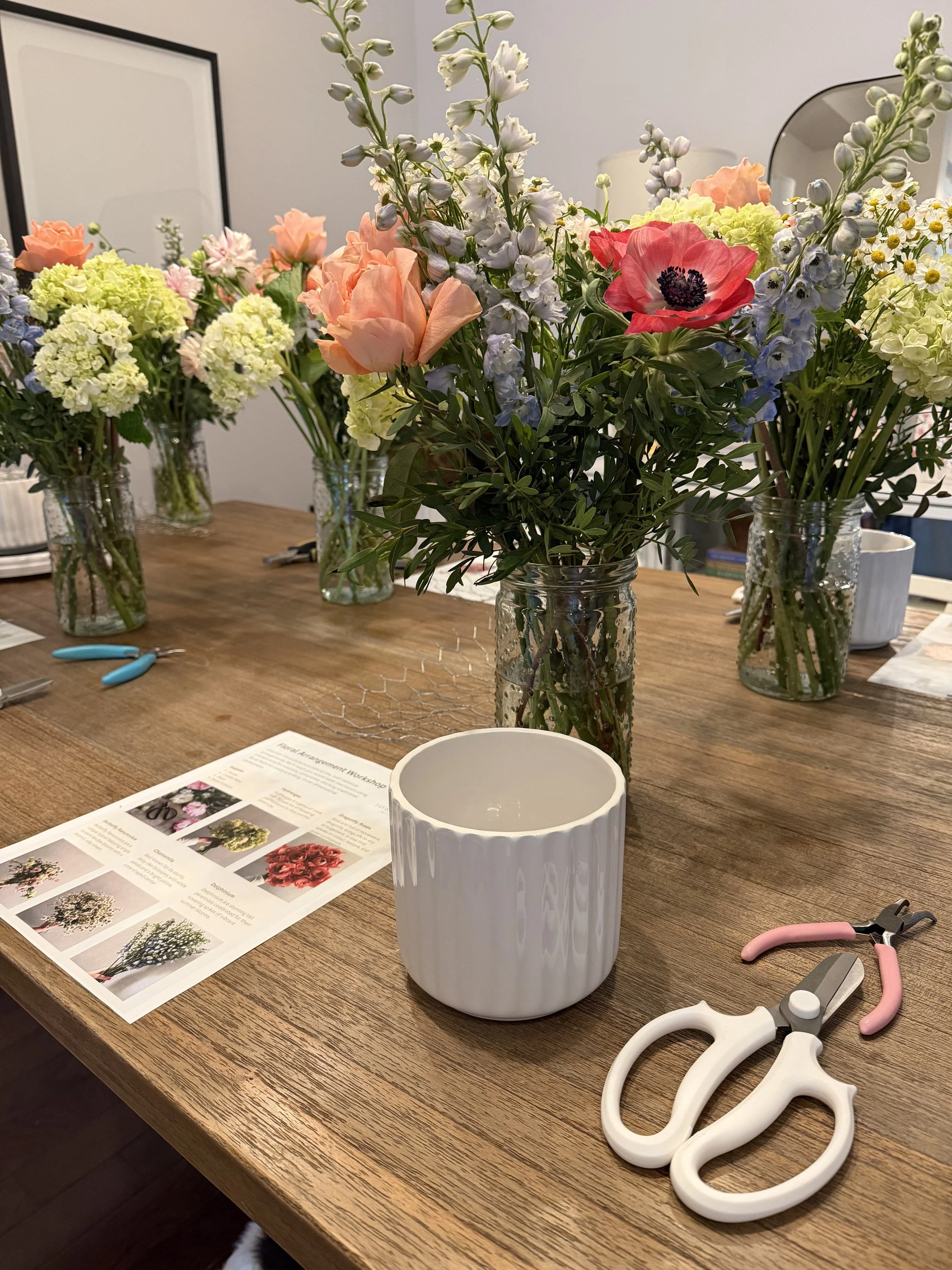 A wooden table with various flower arrangements in glass vases, a white ceramic pot, and flower arranging tools like wire cutters and floral scissors. A printed guide about flower arrangements is also on the table.