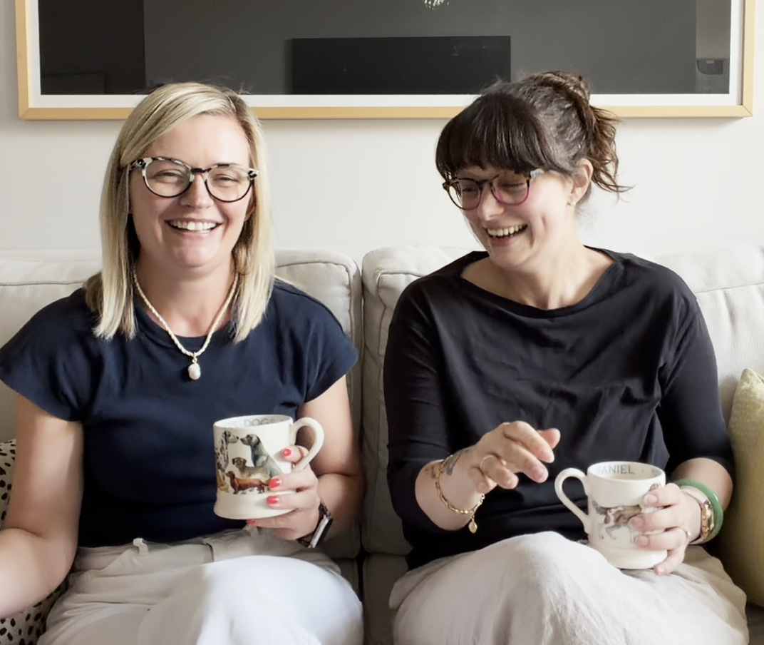 Two women sitting on a sofa, smiling and holding coffee mugs, wearing glasses and casual clothing.