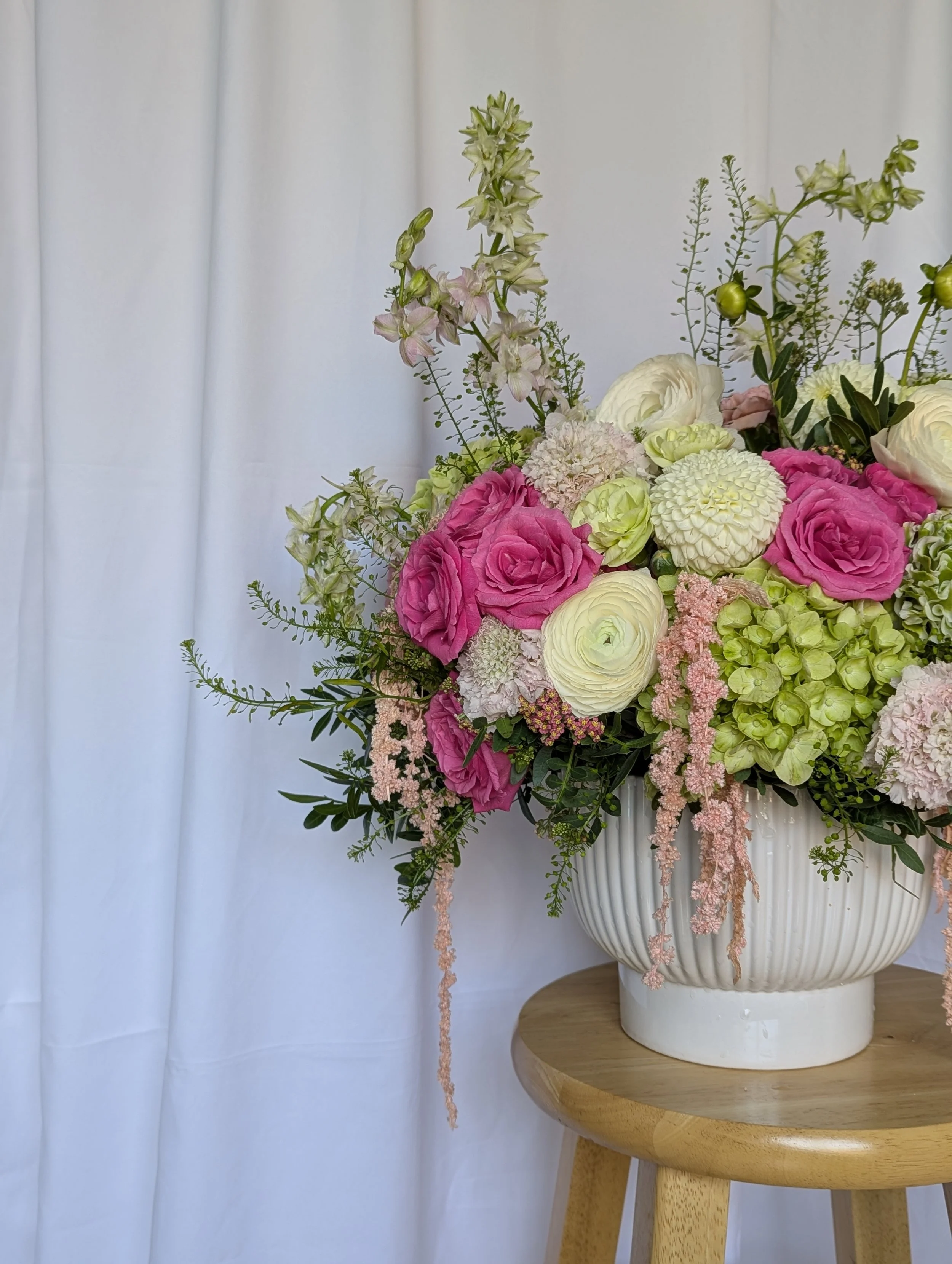 A colorful flower arrangement in a white vase on a small wooden table, with a white curtain in the background.