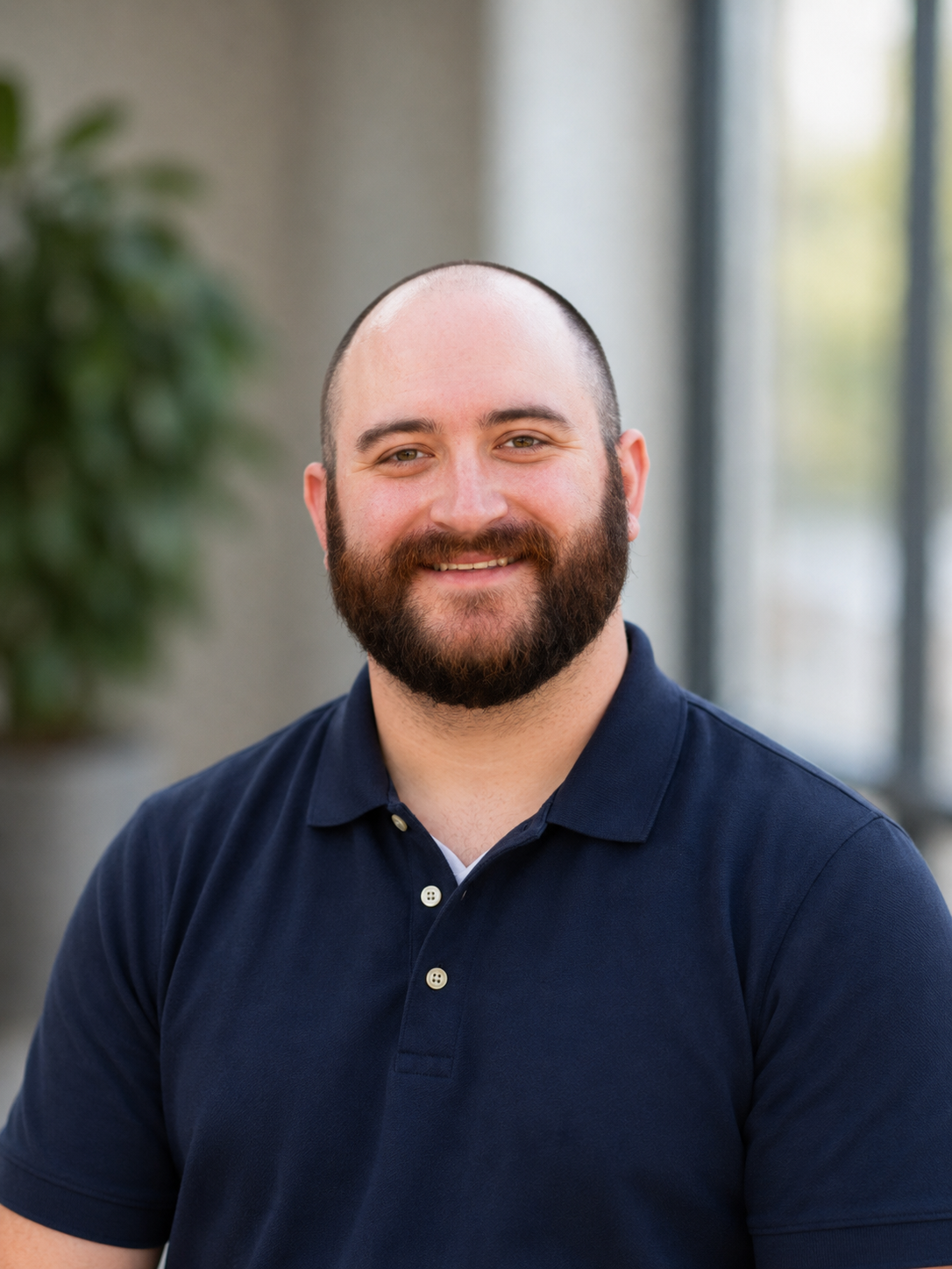 A smiling man with a beard and short hair, wearing a navy blue polo shirt, standing indoors near a window with natural light.