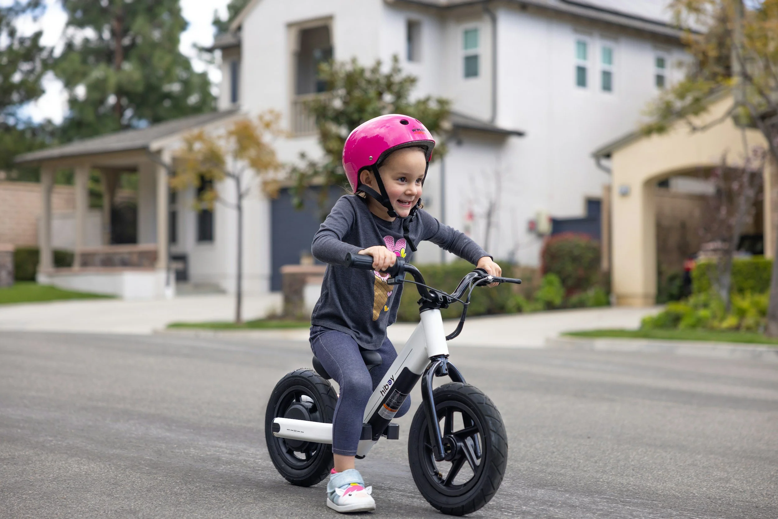 A young girl riding a white bicycle with training wheels on a residential street. She is wearing a pink helmet, a gray long-sleeve shirt with a heart and unicorn design, and gray pants. She appears happy and focused, with trees and houses visible in the background.