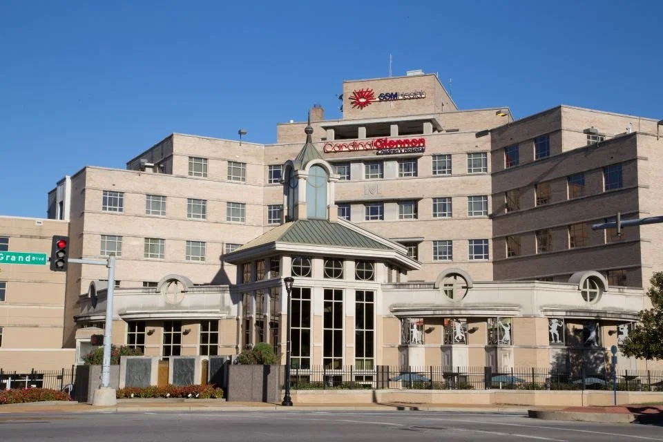 A large beige hospital building with multiple floors, a main entrance with a glass enclosed porch, several windows, and signs for SSM Health and Cardinal Glennon Children's Hospital. Street and traffic lights are in the foreground.