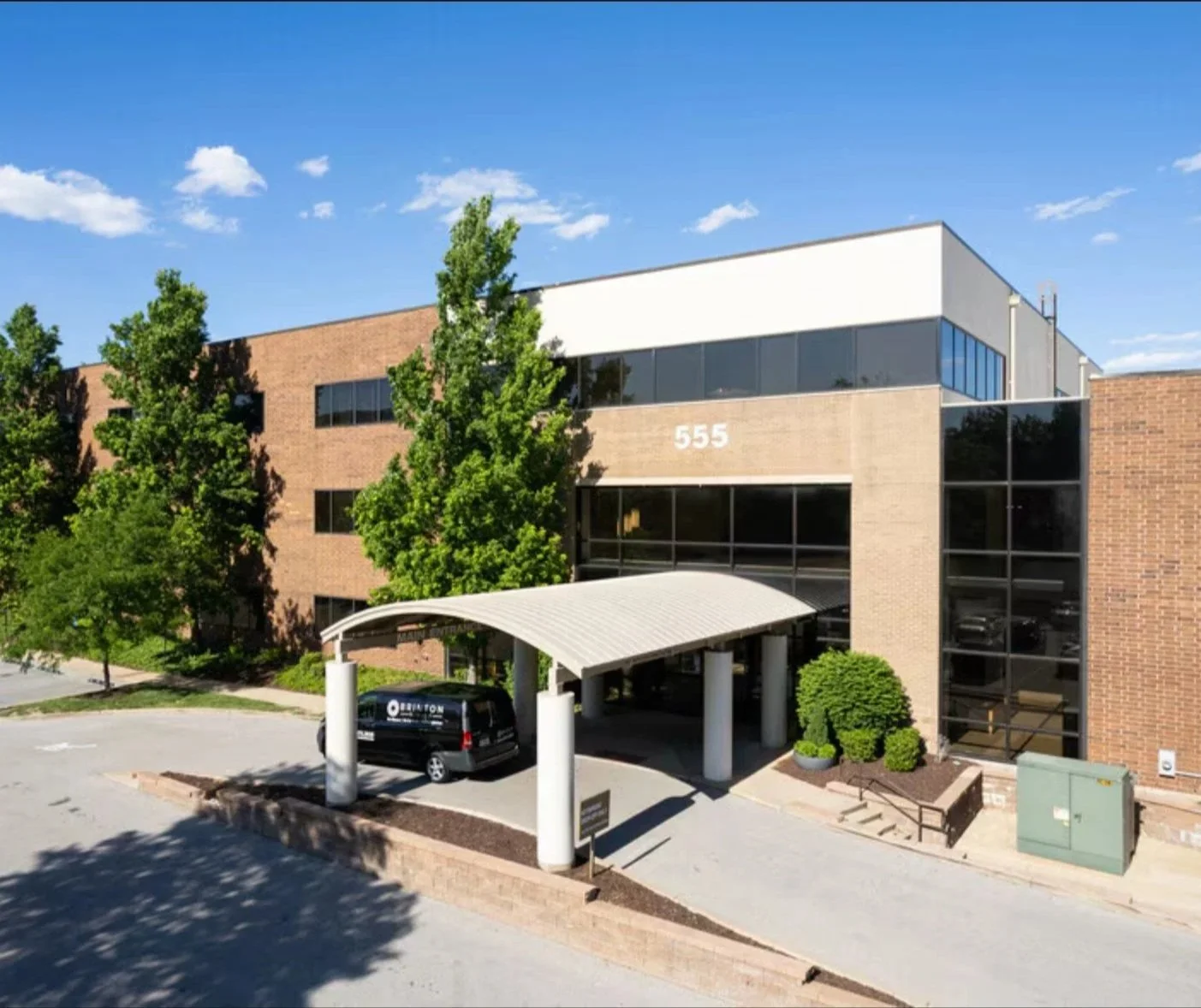 Photo of a modern three-story office building with brick and glass exterior, site number 555, surrounded by trees and featuring a covered entrance with a parked black van.