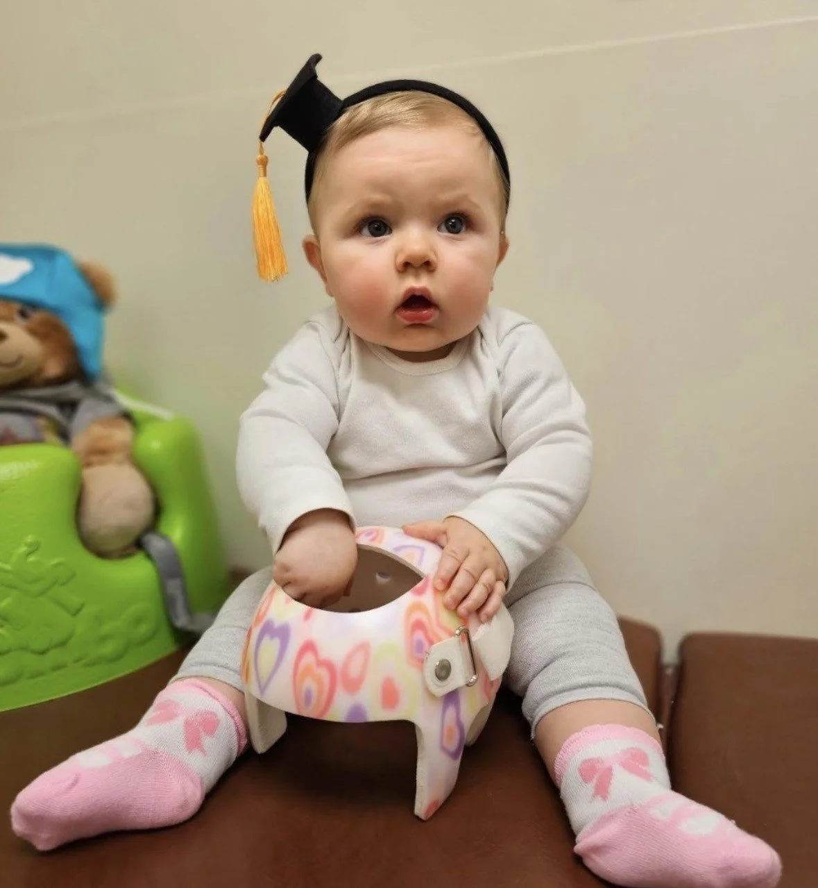 A baby girl sitting on a brown surface wearing a graduation cap, a white long sleeve shirt, gray pants, and pink socks with bows, holding a cranial remolding helmet with colorful heart patterns.