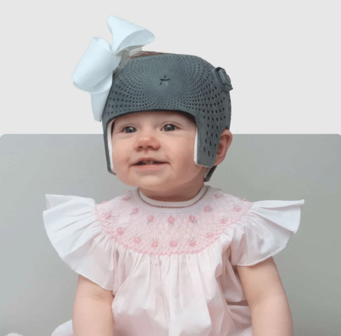 A smiling baby girl wearing a dark gray 3D cranial remolding helmet with a white bow on top and a white smocked dress with ruffled sleeves.