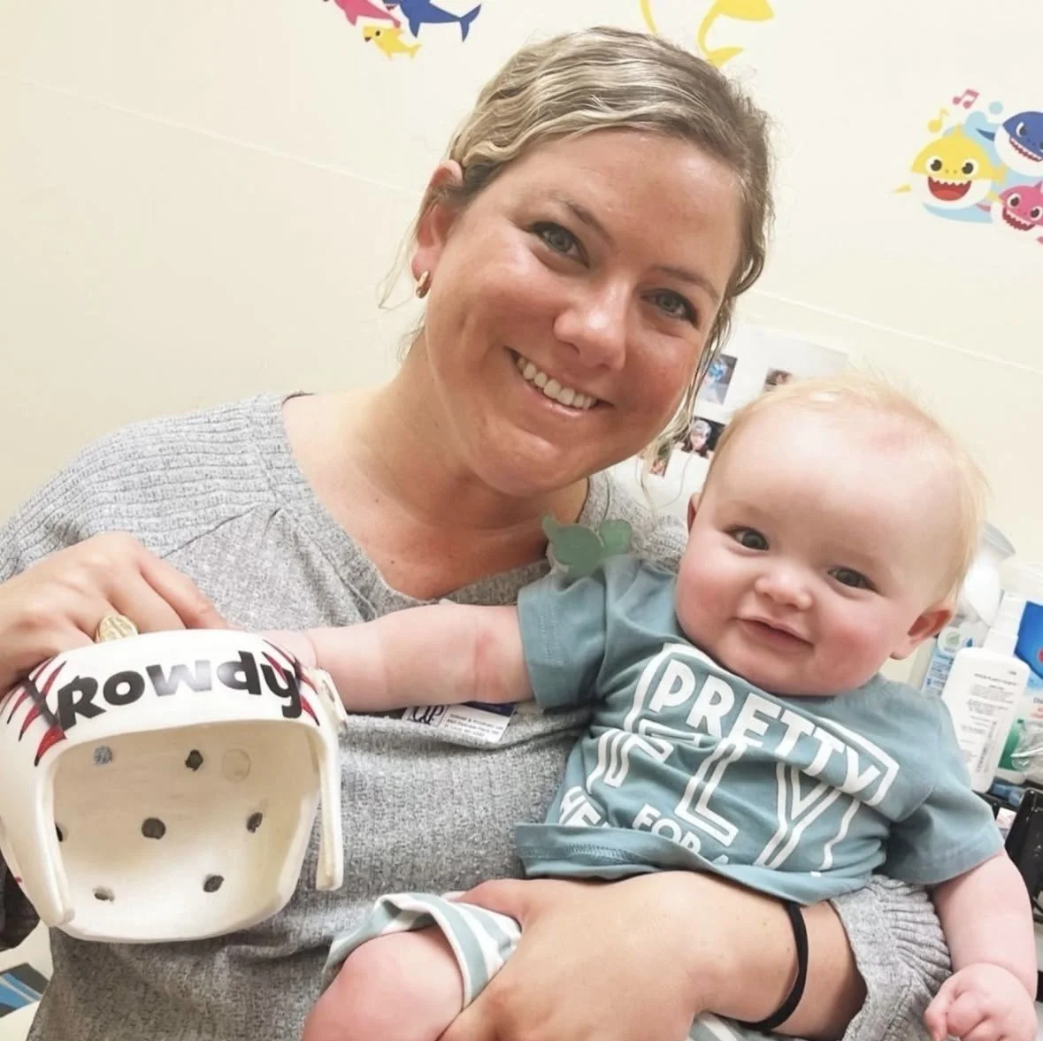 A woman smiling and holding a happy baby with a cranial remolding helmet in his hands.