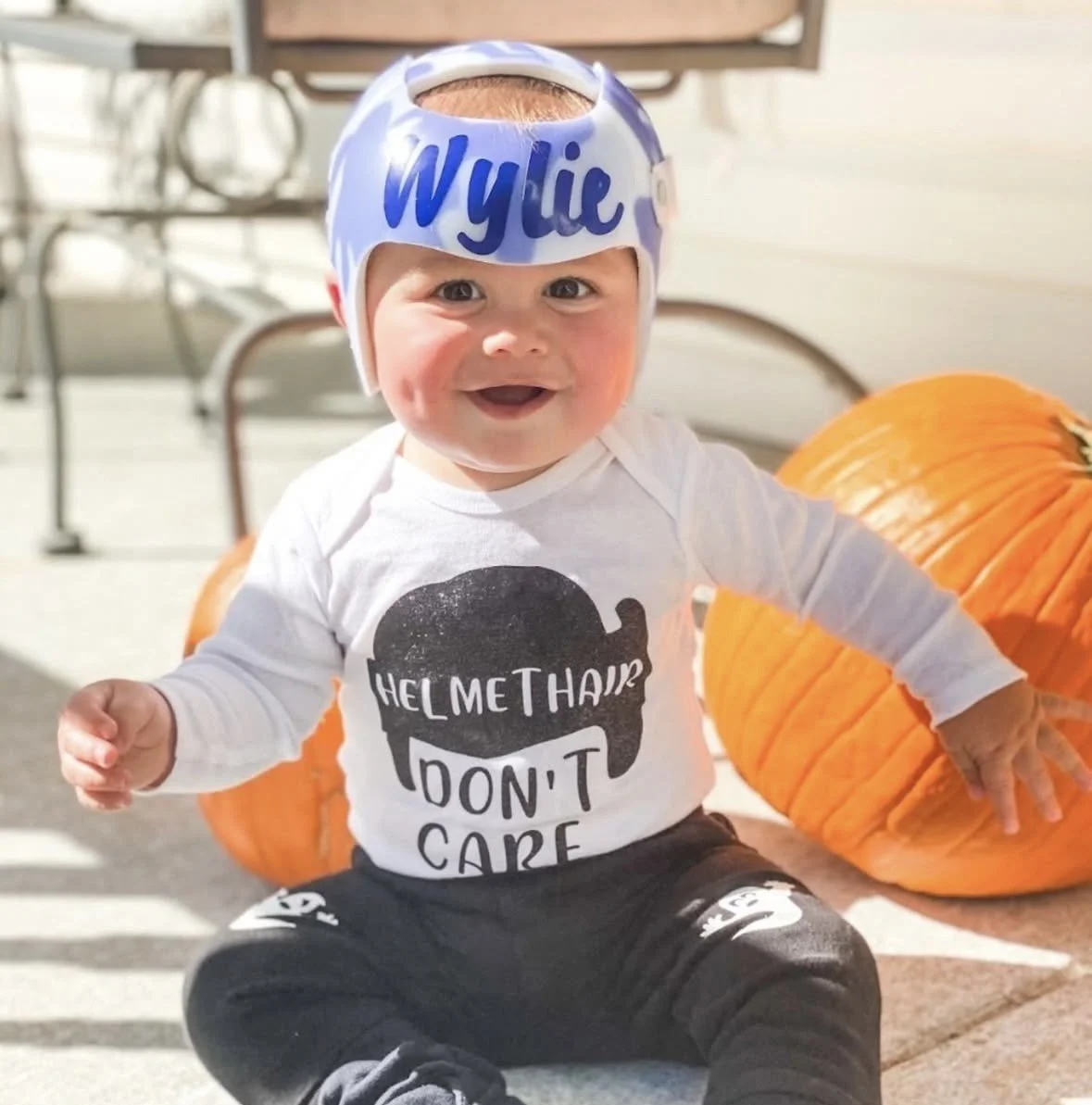 A smiling baby sitting on the ground with two pumpkins behind him, wearing a helmet labeled "Wylie" and a shirt that says "Helmet hair I don't care."