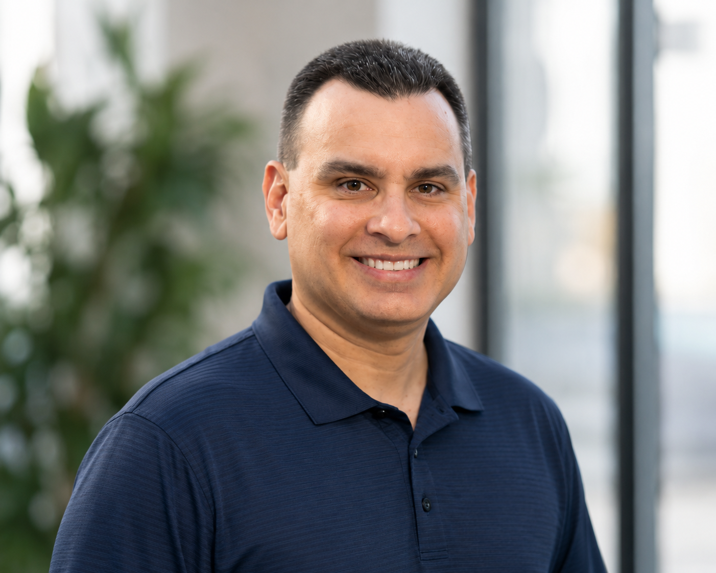 A man with short dark hair and light skin, smiling at the camera, wearing a navy blue collared shirt, in an indoor setting with large windows and a blurred green plant in the background.
