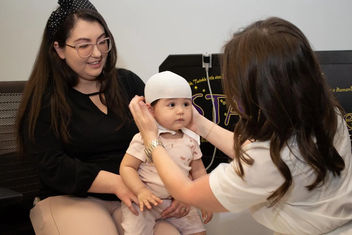 A young woman with long brown hair and glasses, smiling, sitting on a chair while a baby in a white cap is being examined by a healthcare professional. The woman holds the baby, and the professional is adjusting the baby's cap.