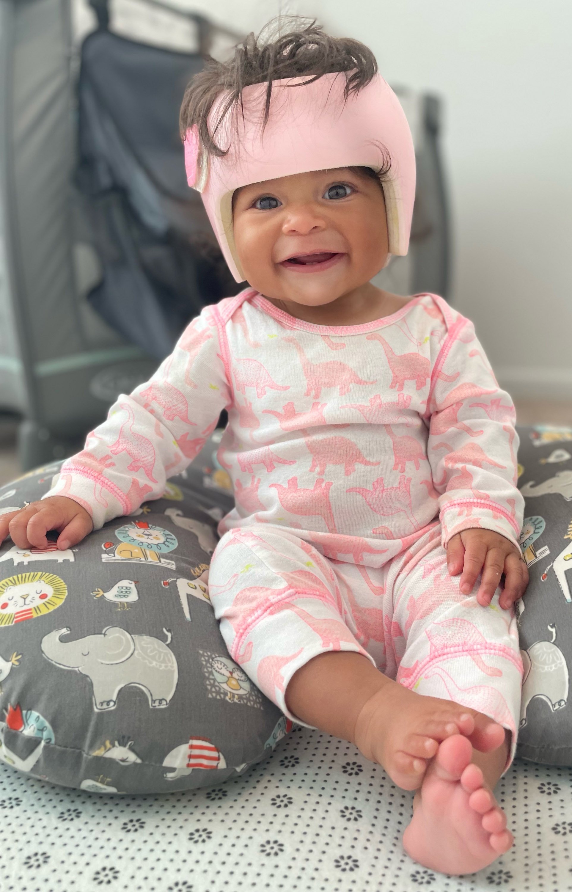 A smiling baby girl with dark hair wearing a pink cranial remolding helmet and pajamas with pink and yellow dinosaurs, sitting in a baby chair with a patterned cushion, holding onto her foot.
