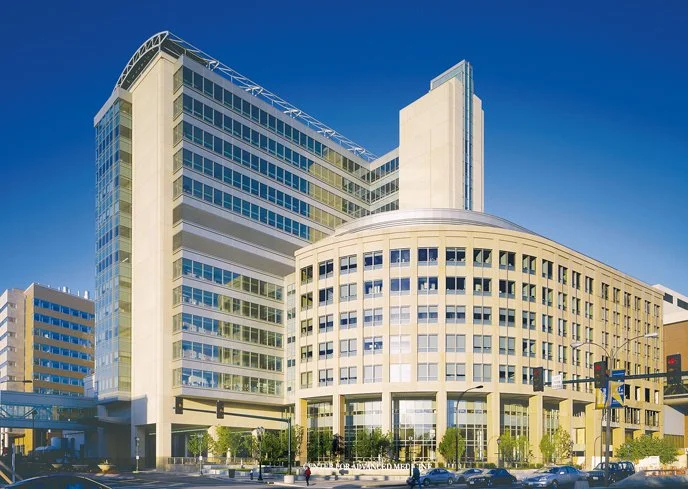 Modern multi-story office building with glass windows and concrete exterior in an urban setting, under a clear blue sky.