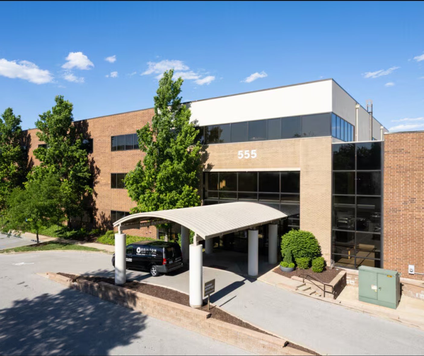 A modern office building with a brick exterior, large glass windows, and the number 555 displayed on the front. There are trees, a black van parked under a white canopy, and a clear blue sky with scattered clouds.