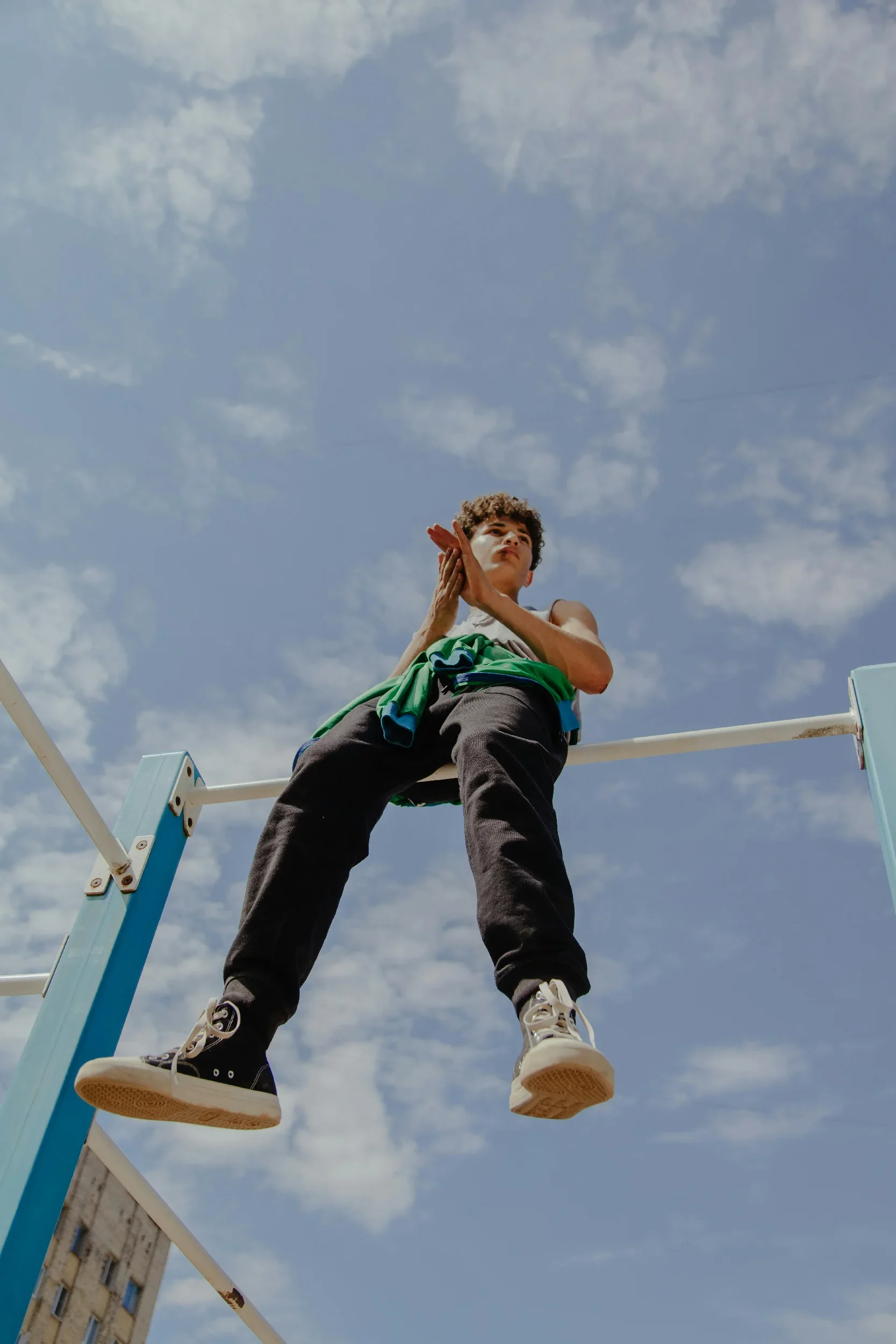 A young person standing on a ladder outdoors under a partly cloudy sky, looking upwards with hands clasped together.