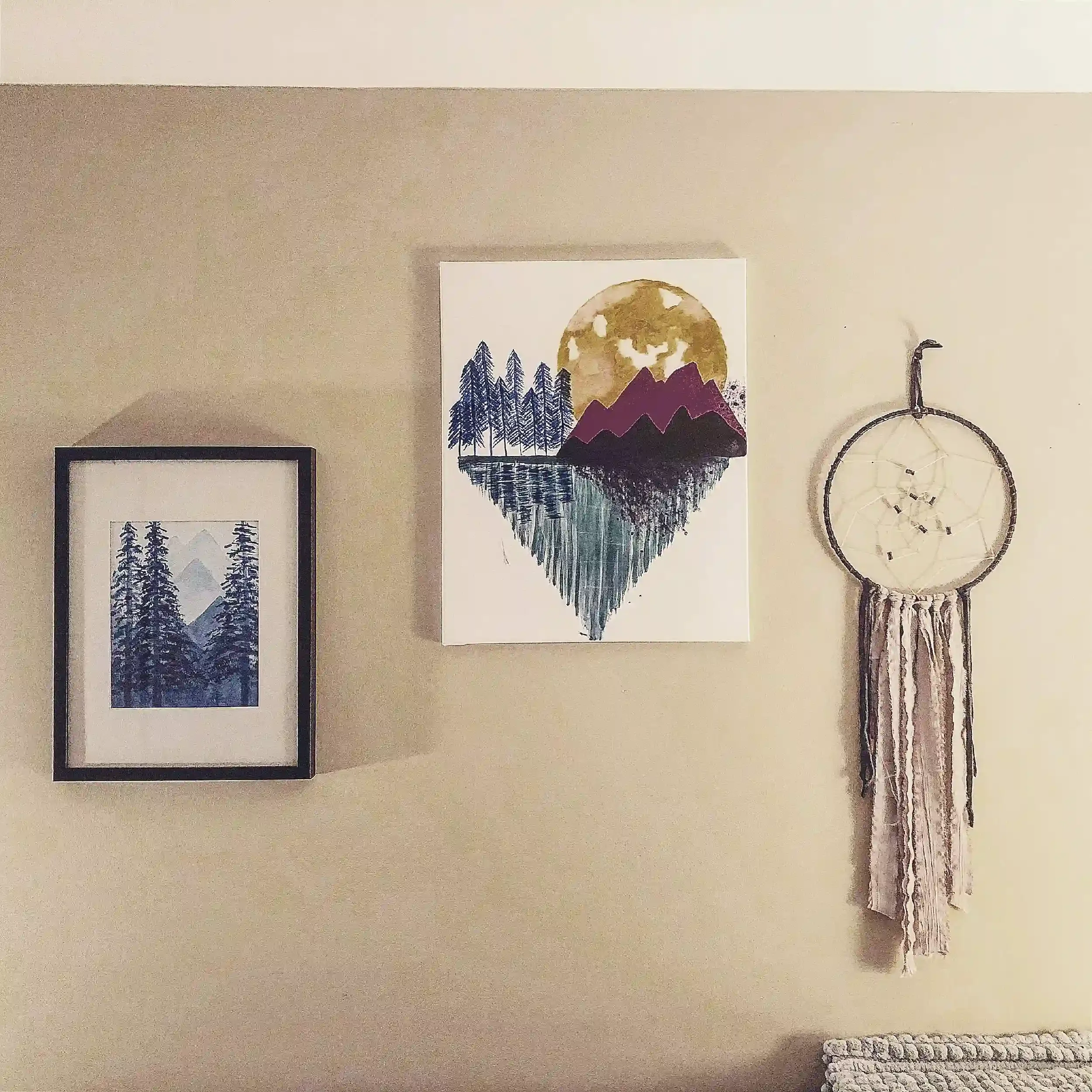 Decorative wall with two framed prints of mountain scenes, a large round watercolor artwork of mountains and a sun, a dreamcatcher, and a woven basket.