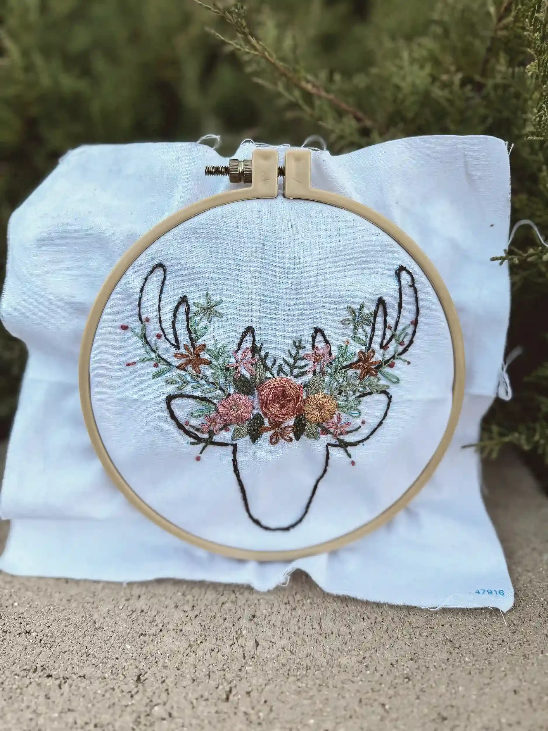 Embroidery hoop with floral embroidered design on white fabric, placed on ground with green foliage in the background.