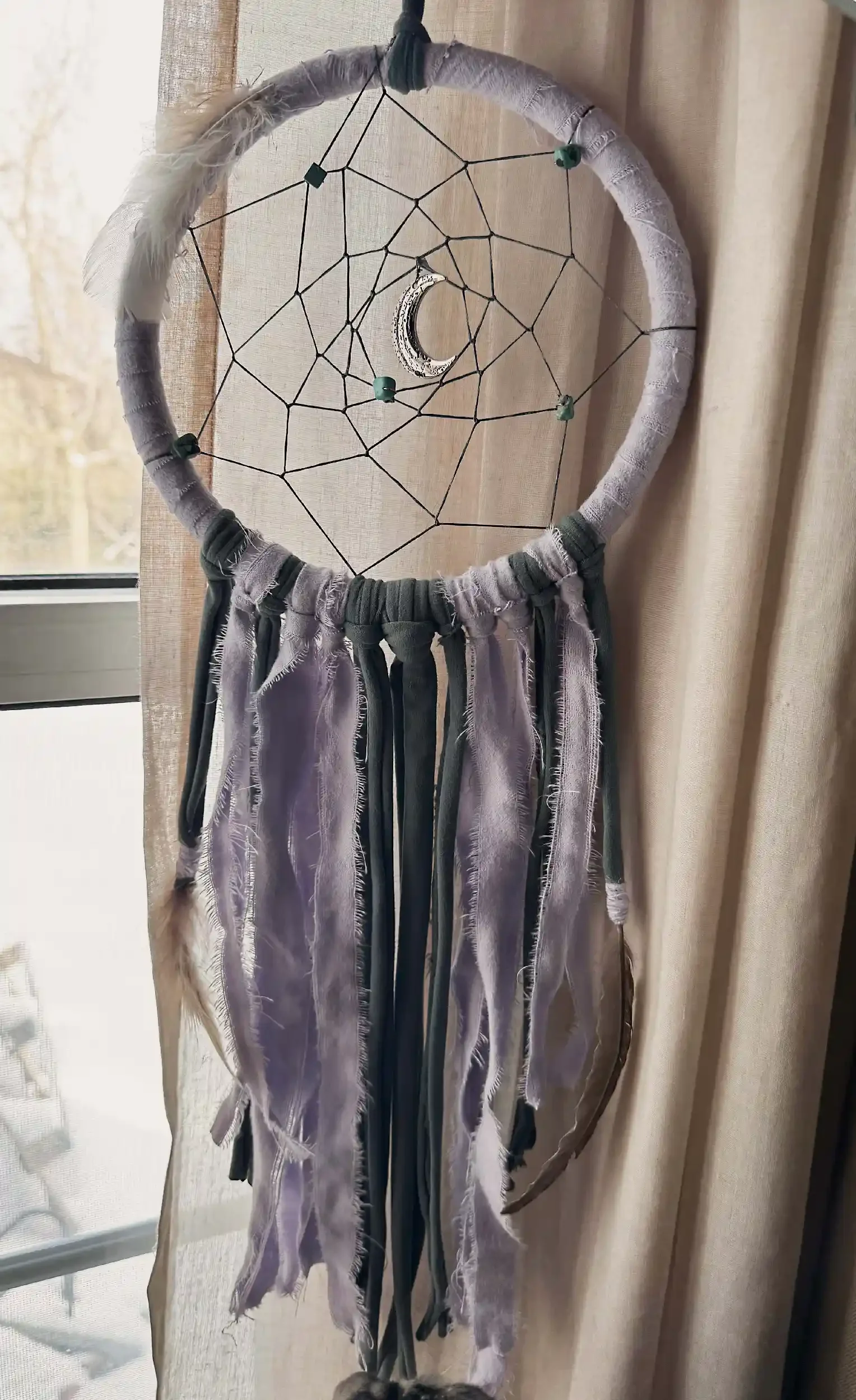 A dreamcatcher with a black circular frame wrapped in fabric, featuring a crescent moon in the center and a web pattern with beads, decorated with long, tattered fabric strips hanging down.