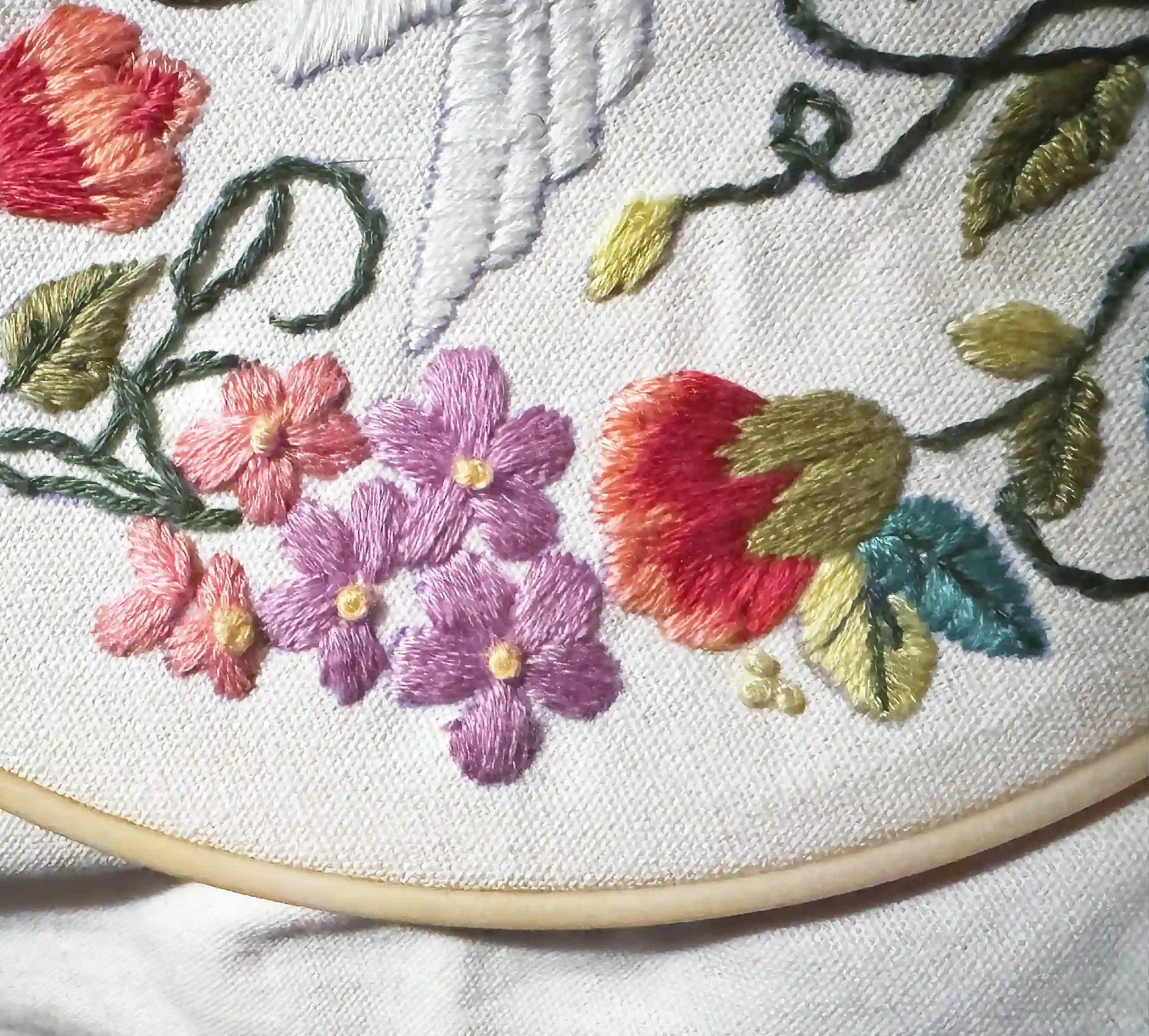 Embroidery of colorful flowers and green leaves on white fabric, framed with a wooden hoop.