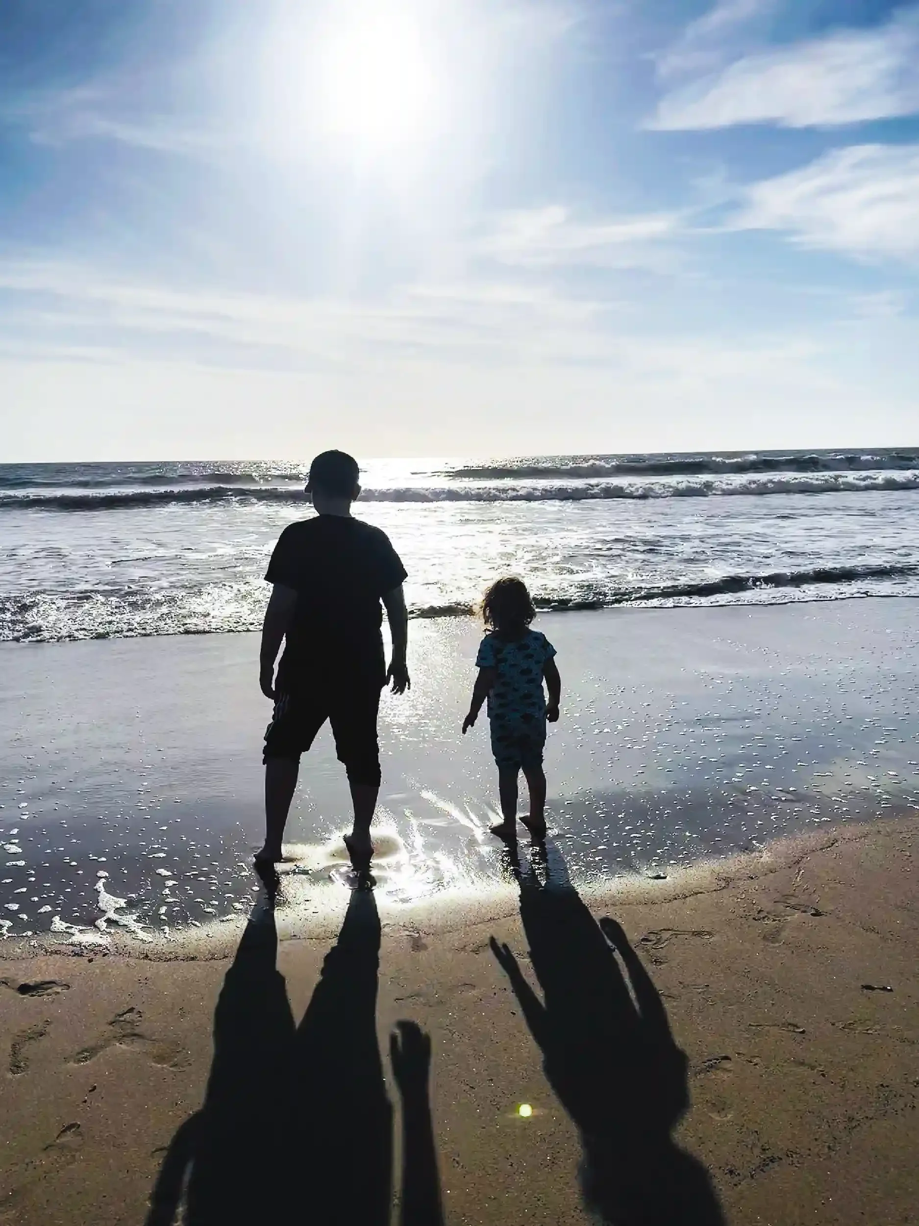 Two children, one adult, and a dog walking along the beach at sunset or sunrise with the ocean waves in the background and the sun shining brightly in the sky.