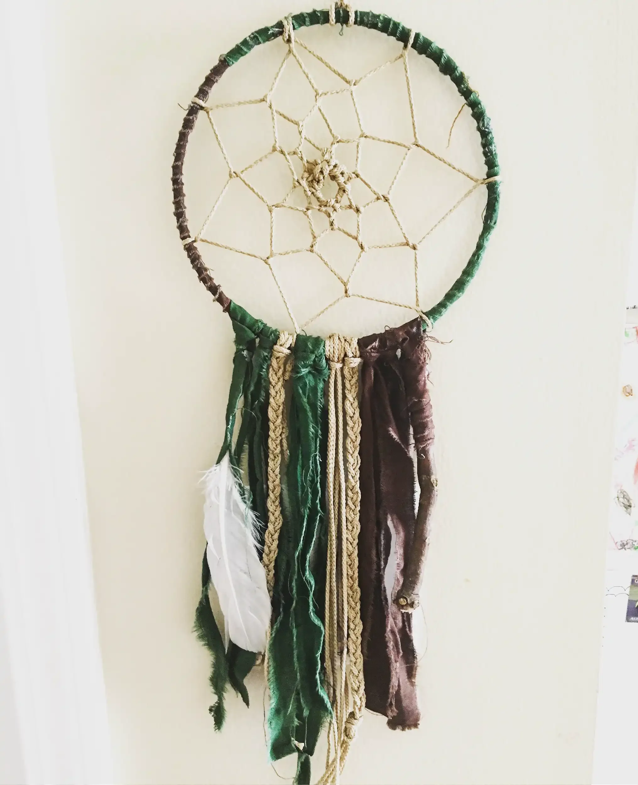 A dreamcatcher made of a metal or plastic hoop wrapped in green and brown cloth, with a web string inside and various pieces of cloth or fabric hanging from the bottom.