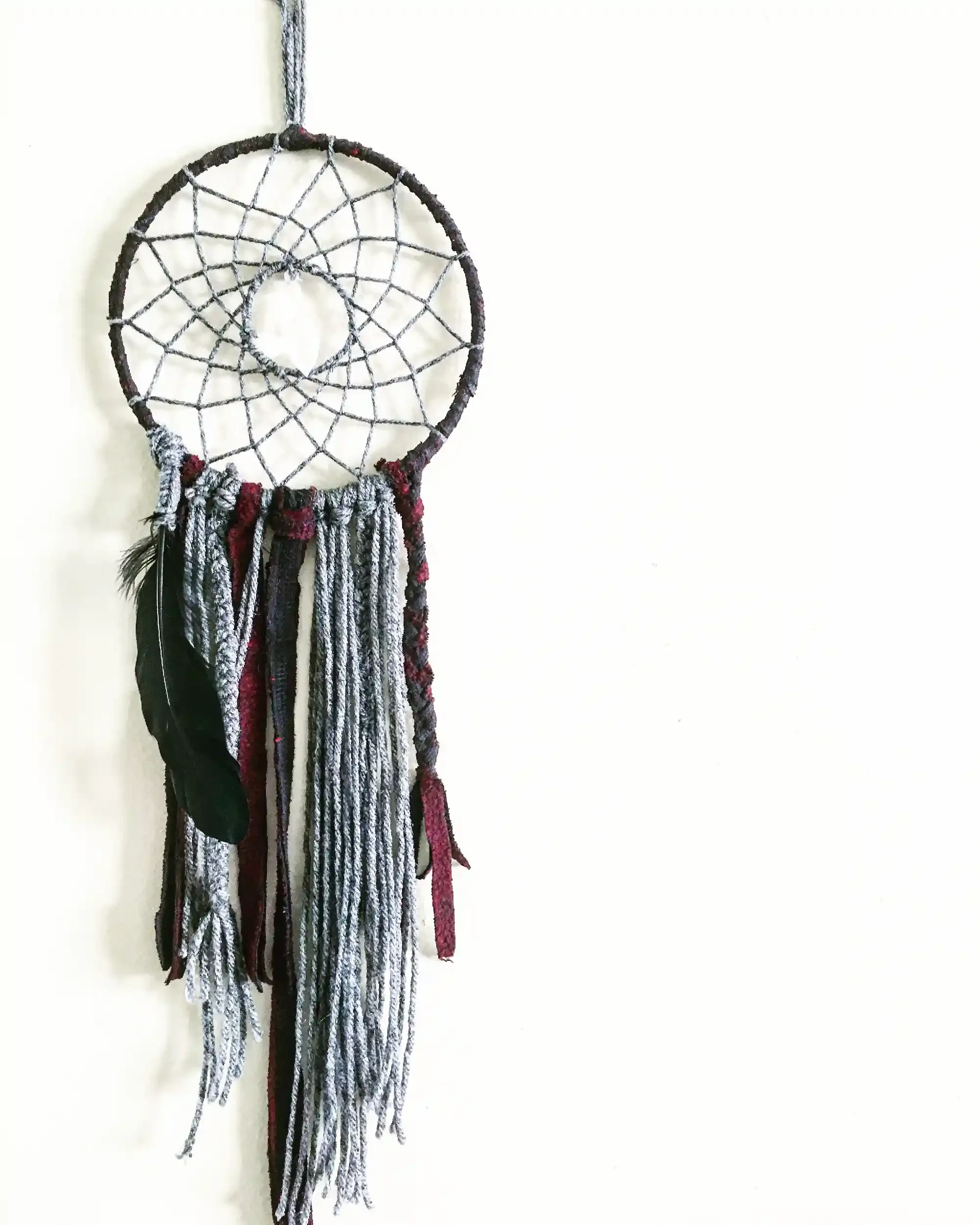 A dreamcatcher hanging on a white wall, with black, red, gray, and white strands of yarn hanging from it.