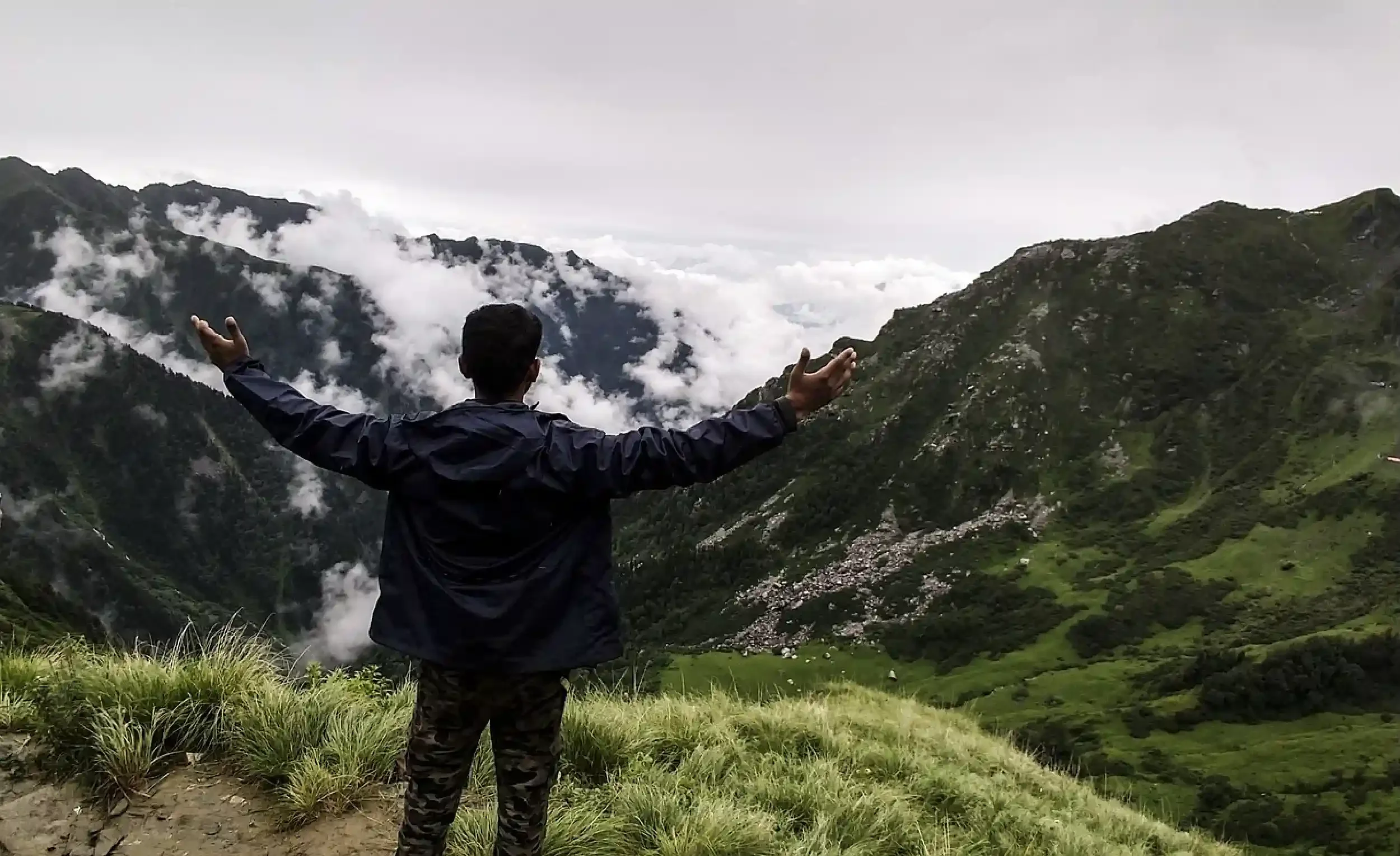 Person with arms outstretched overlooking a mountainous landscape with clouds and greenery.