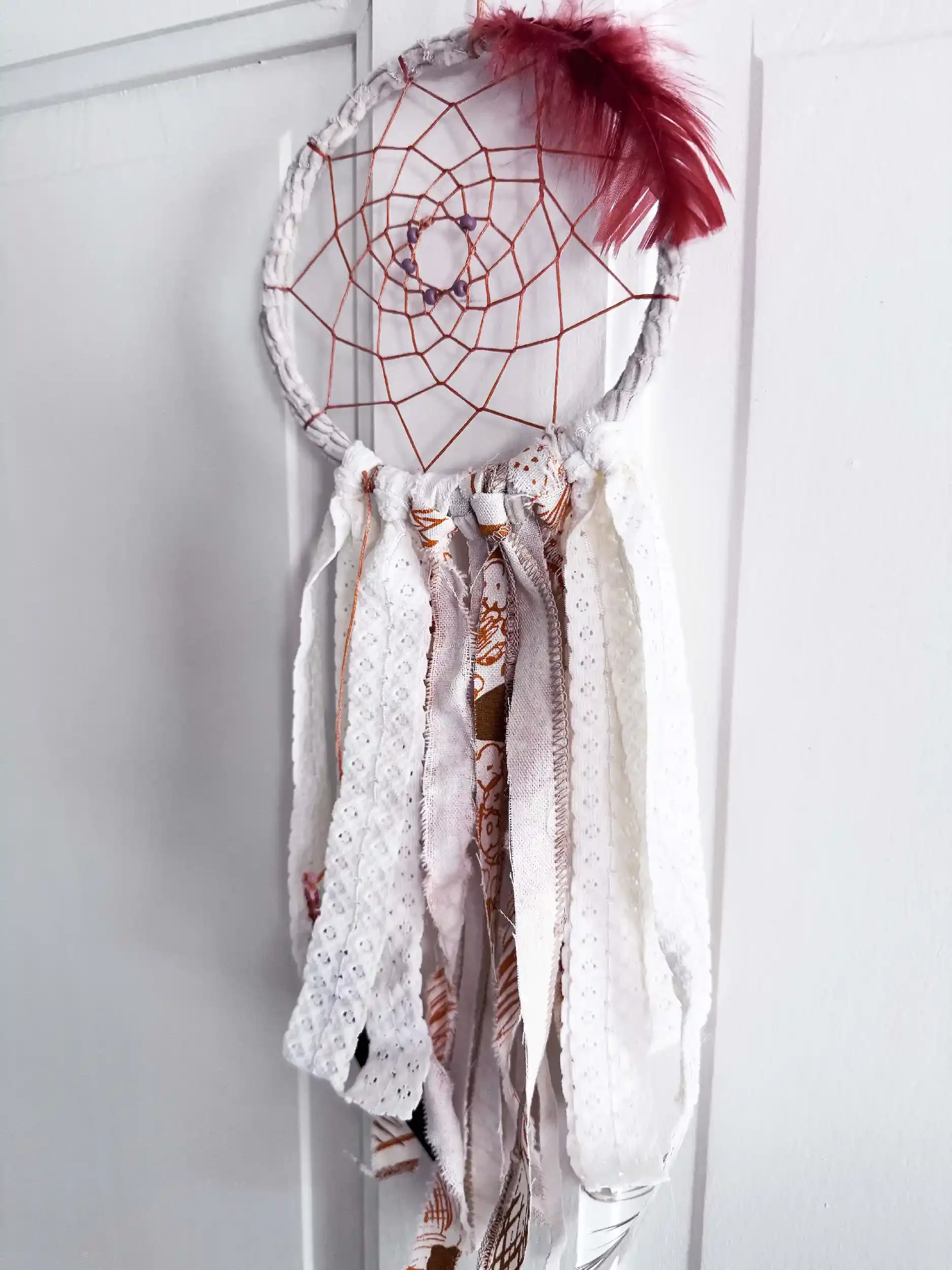 A dreamcatcher with a circular white woven frame, red string web, three purple beads in the center, and red feathers on the top right, hanging against a plain white wall.