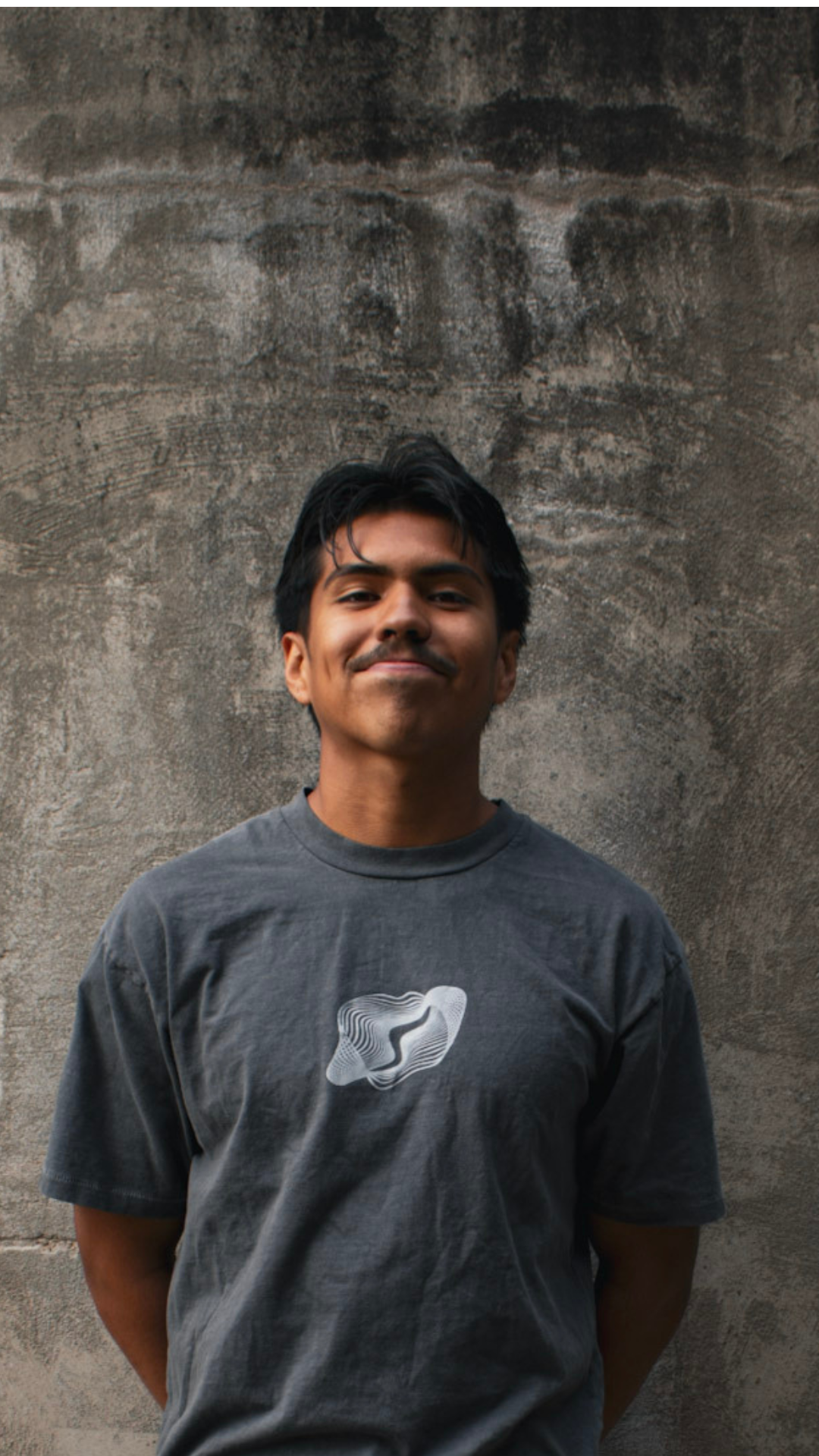 A young man with black hair and a slight smile, standing against a textured concrete wall, wearing a dark gray t-shirt with a white abstract graphic design.