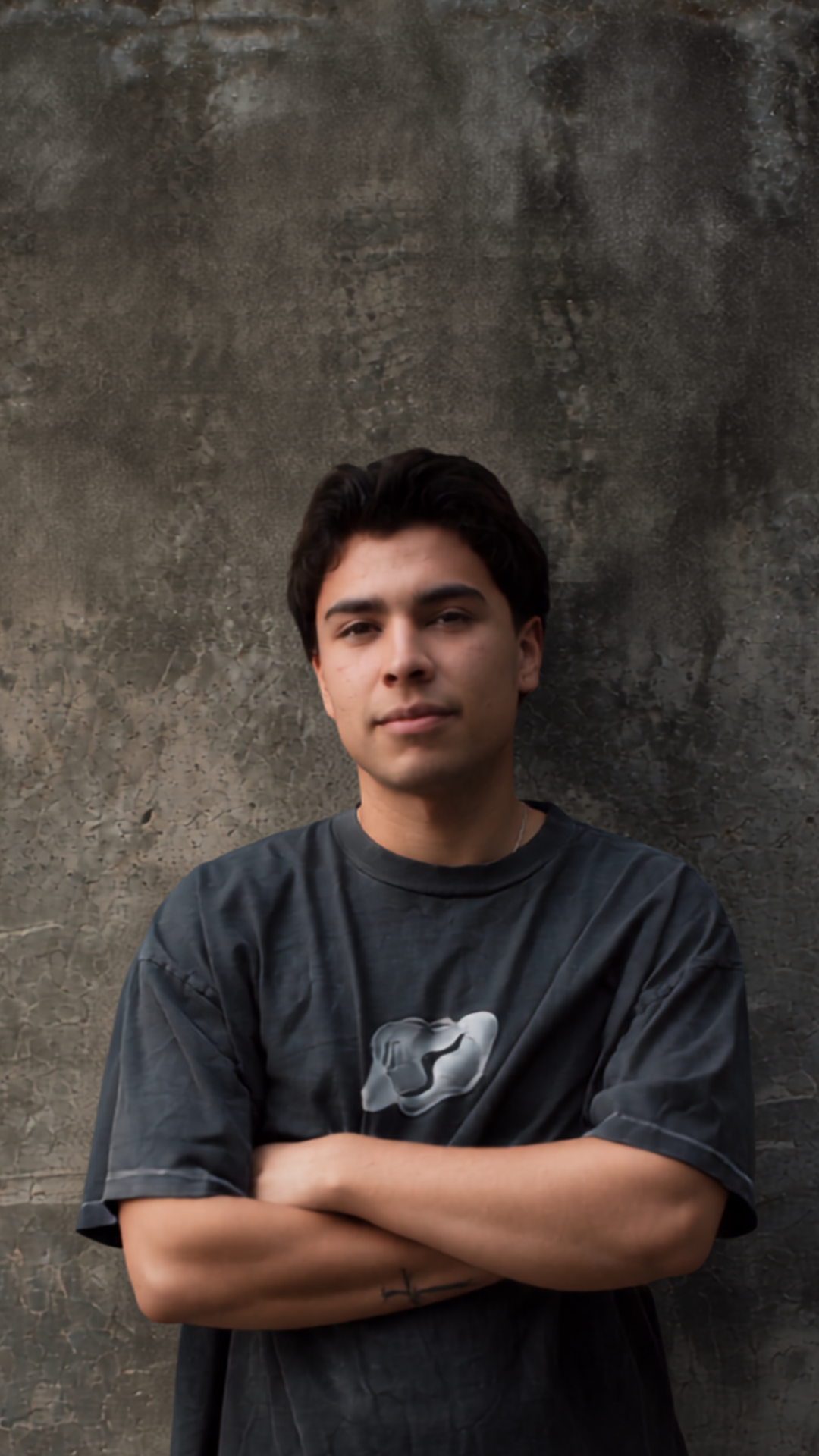 A young man with dark hair and a light complexion standing with arms crossed in front of a textured concrete wall.