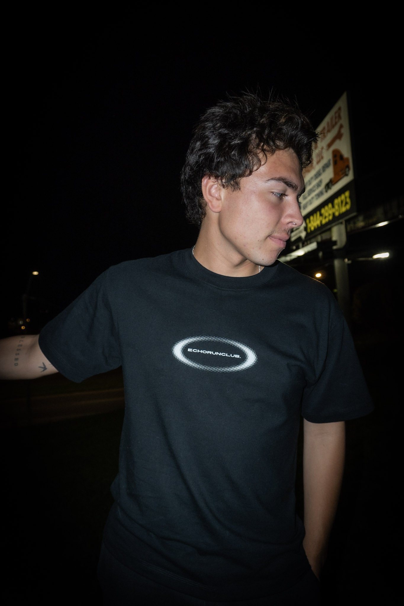 A young man with curly dark hair and light skin wearing a black T-shirt with a circular logo that says "Echorunclub" on it, standing outdoors at night with a billboard in the background.