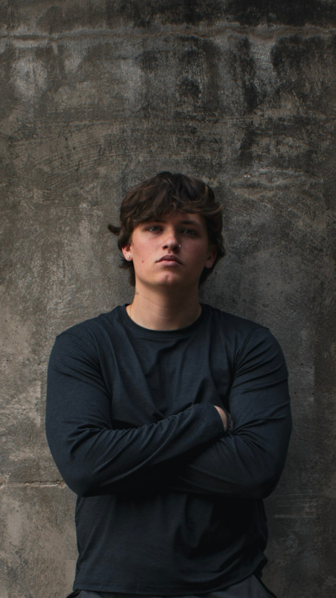 A young man with dark hair, wearing a black long sleeve shirt, standing with folded arms against a textured concrete wall.
