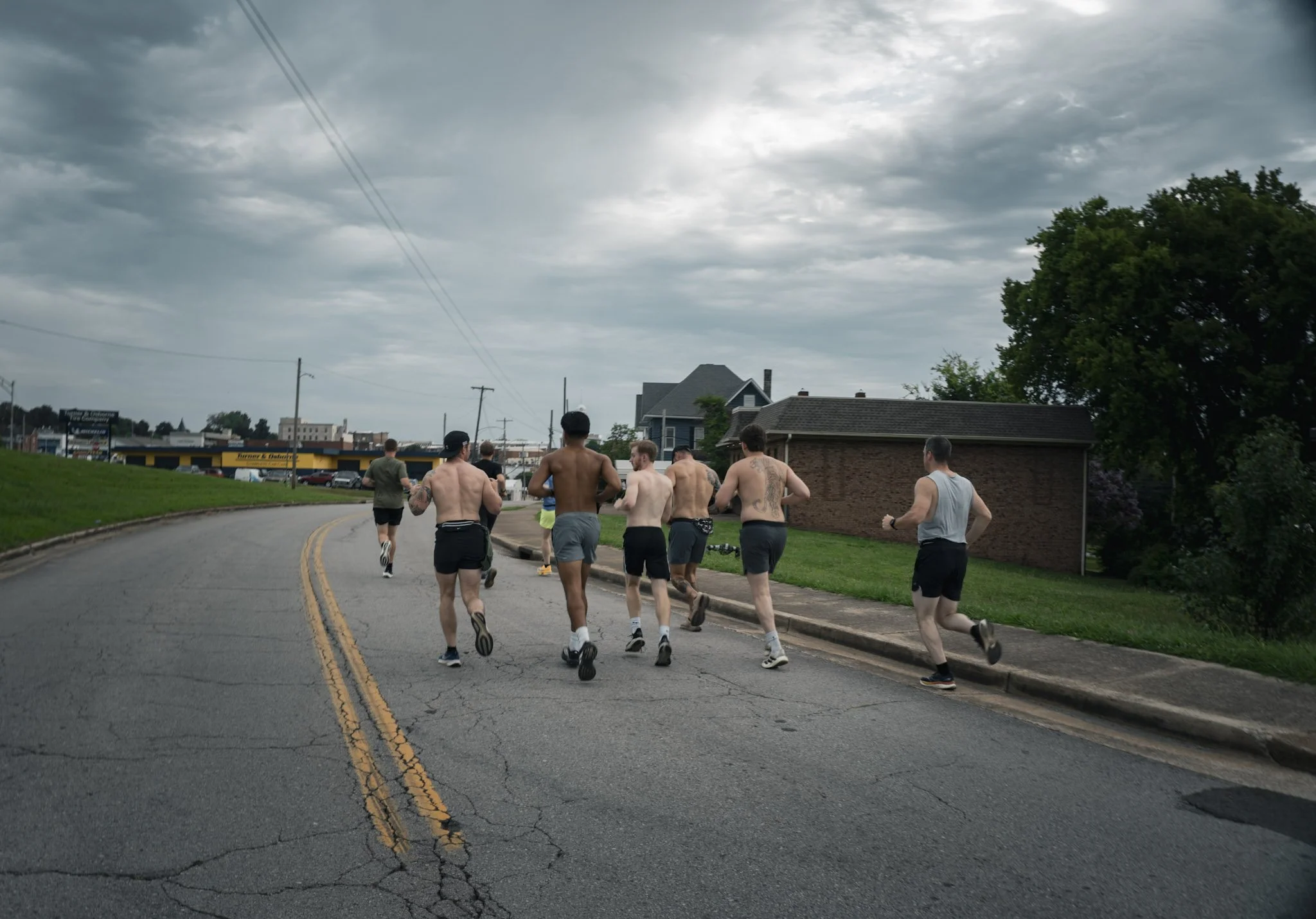 A group of shirtless men running on an urban street under a cloudy sky.