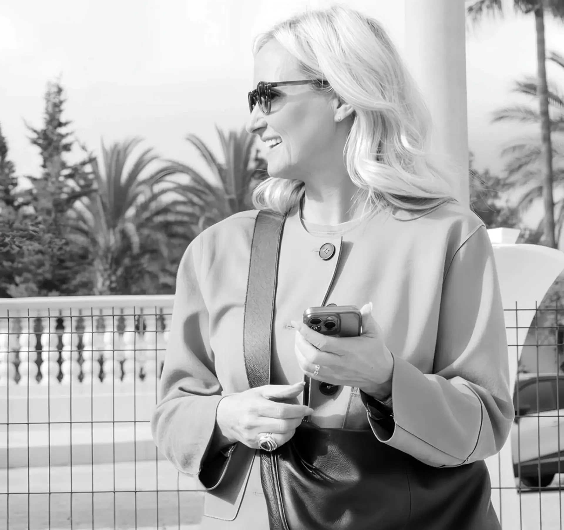 Black and white photo of a woman with blonde hair wearing sunglasses and a diary, standing outdoors near a fence and trees, looking to the side and smiling while holding her phone.