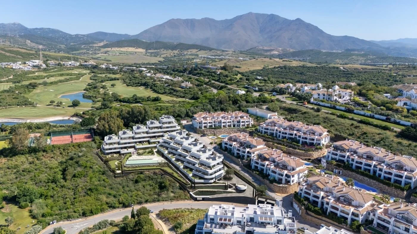 Aerial view of a residential community with white buildings, surrounded by green hills and mountains in the background, featuring sports courts, a pond, and roads.